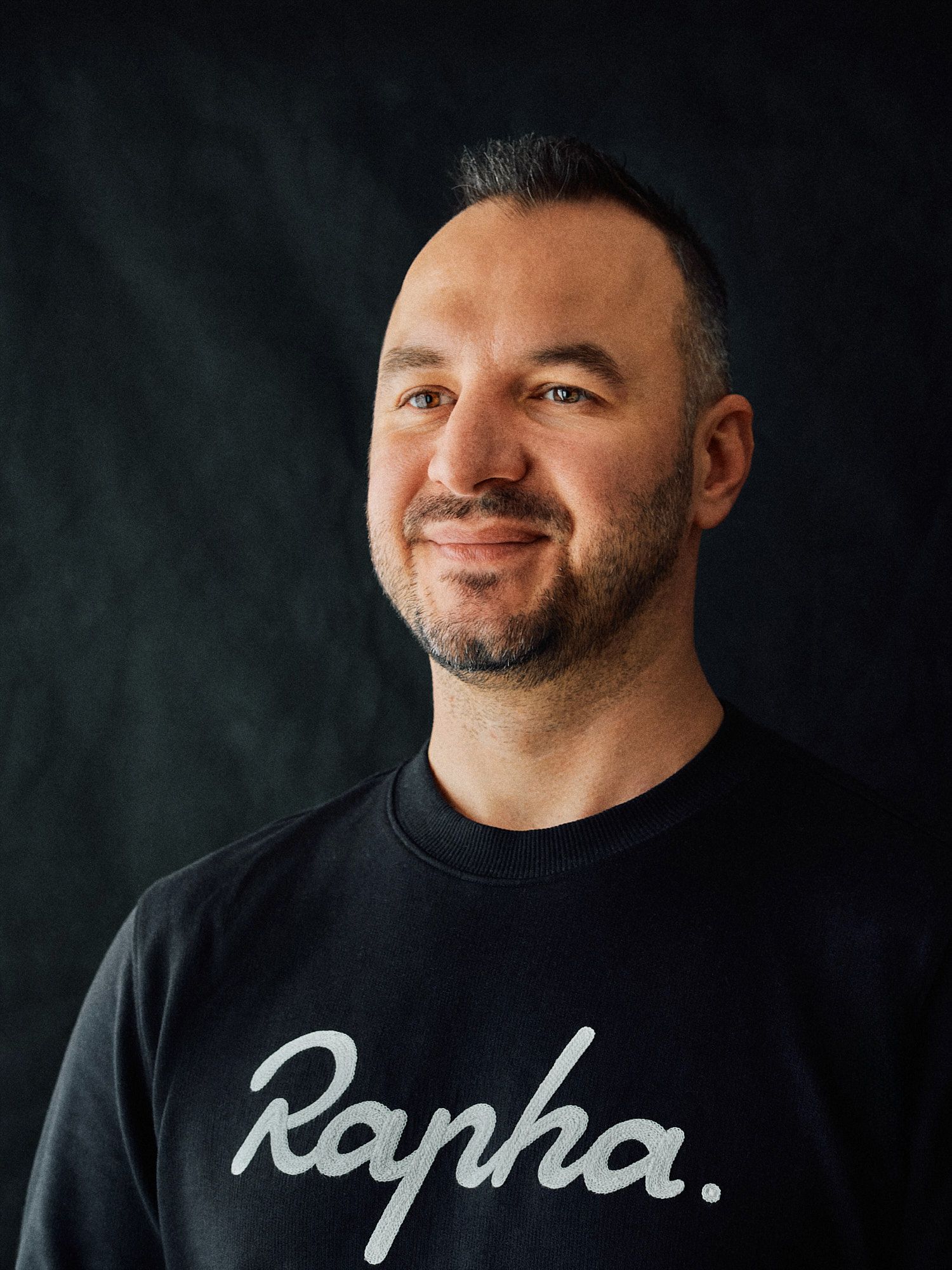 Portrait shot of person in black Rapha sweatshirt smiling against dark backdrop.