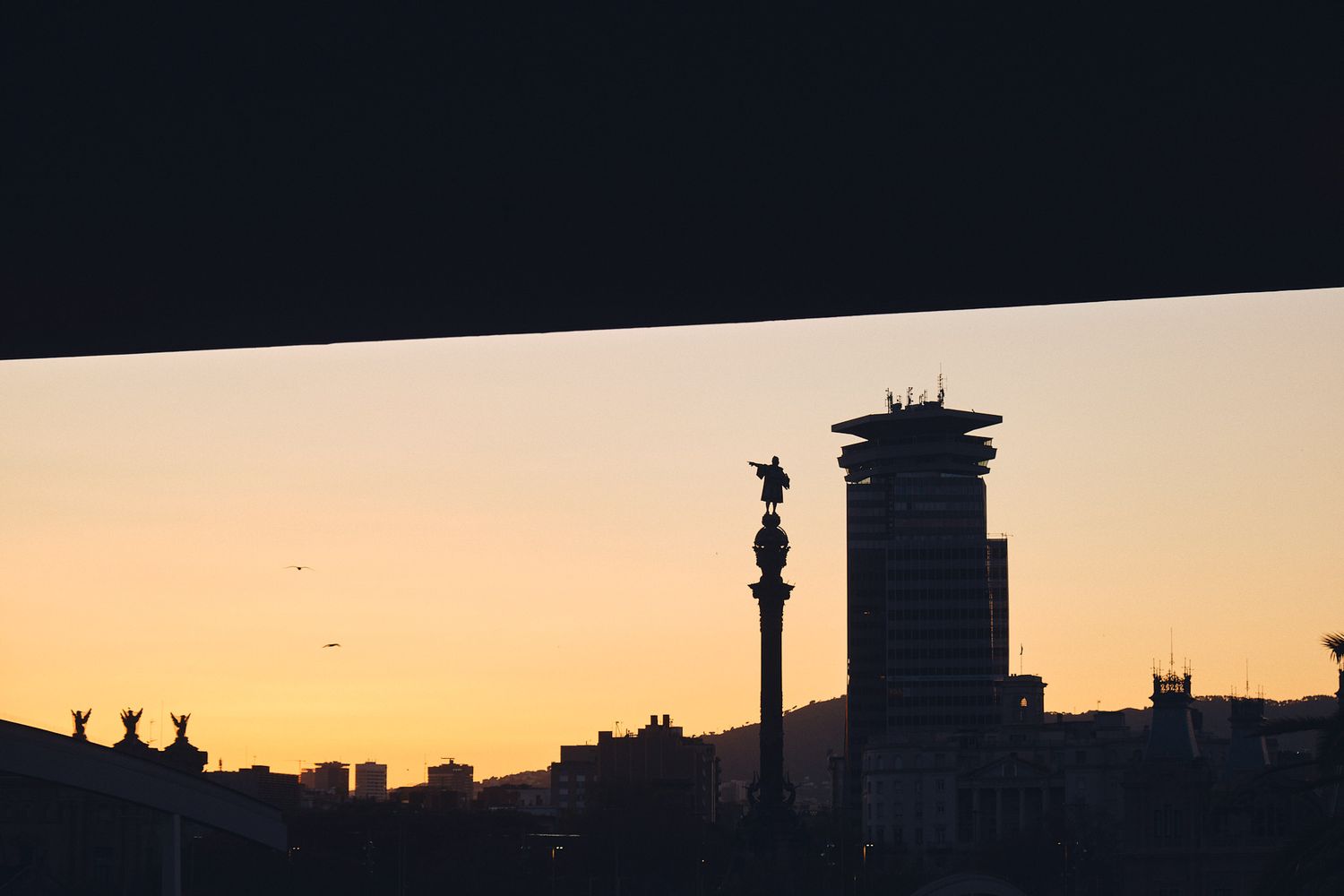 Silhouette of Columbus Monument and city skyline against a golden sunset sky in Barcelona, Spain.