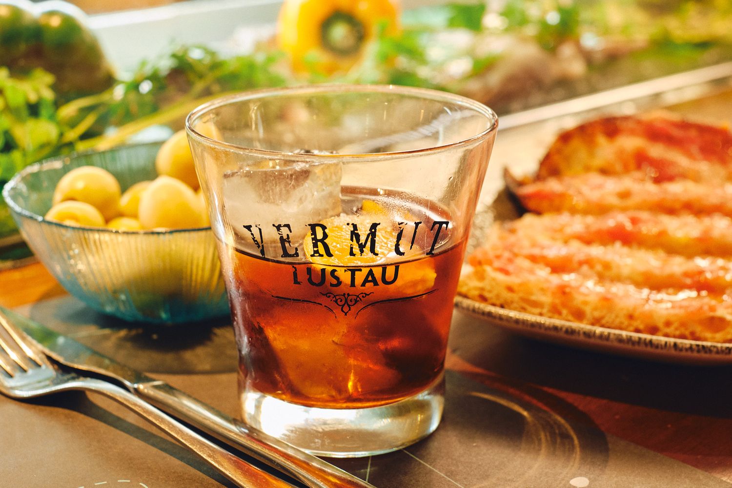A glass of Vermut Lustau served on the rocks, surrounded by appetizers and fresh ingredients on a table.