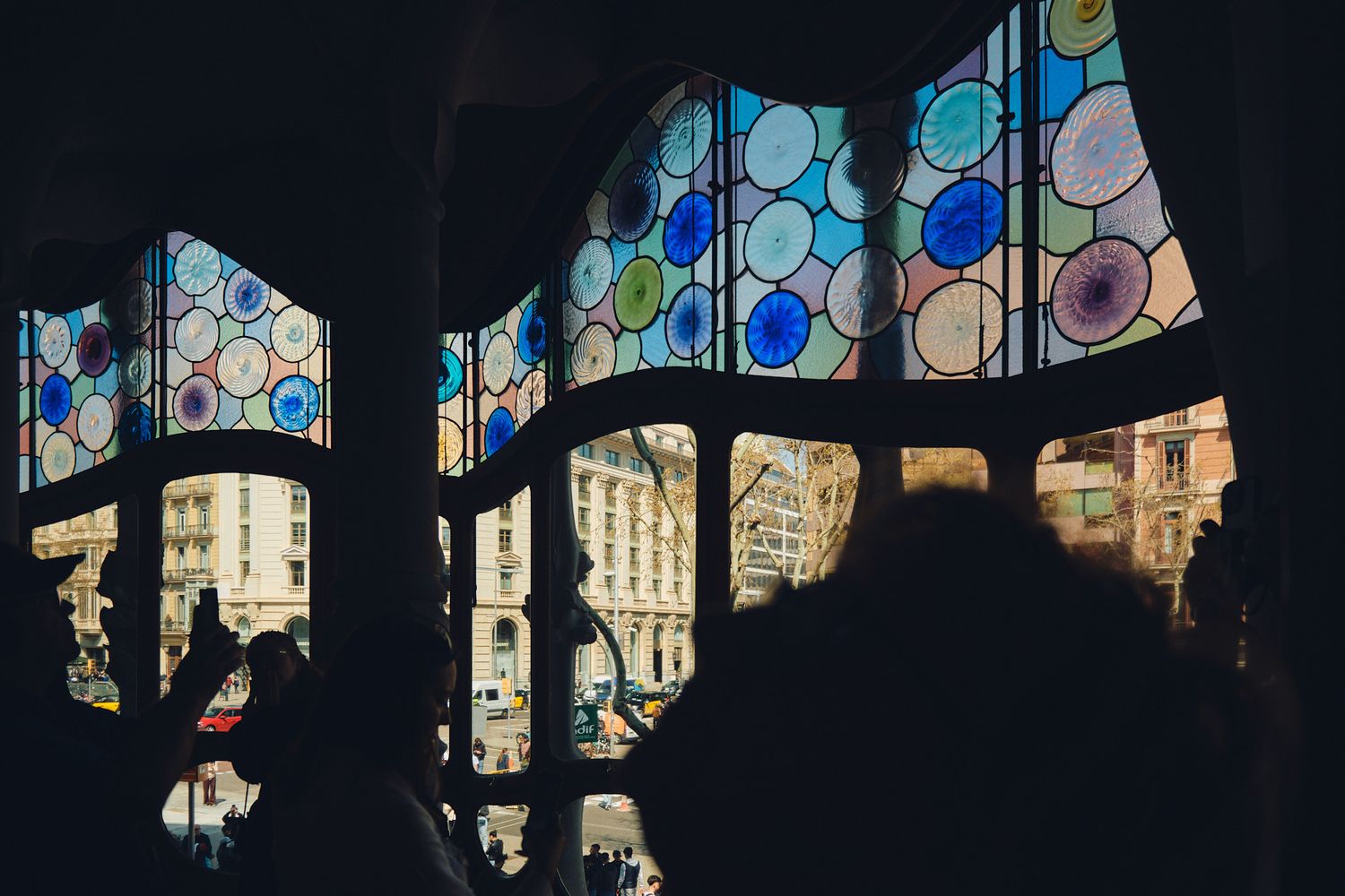 Silhouettes of visitors admire colorful stained glass windows with circular patterns at Casa Batllo in Barcelona.