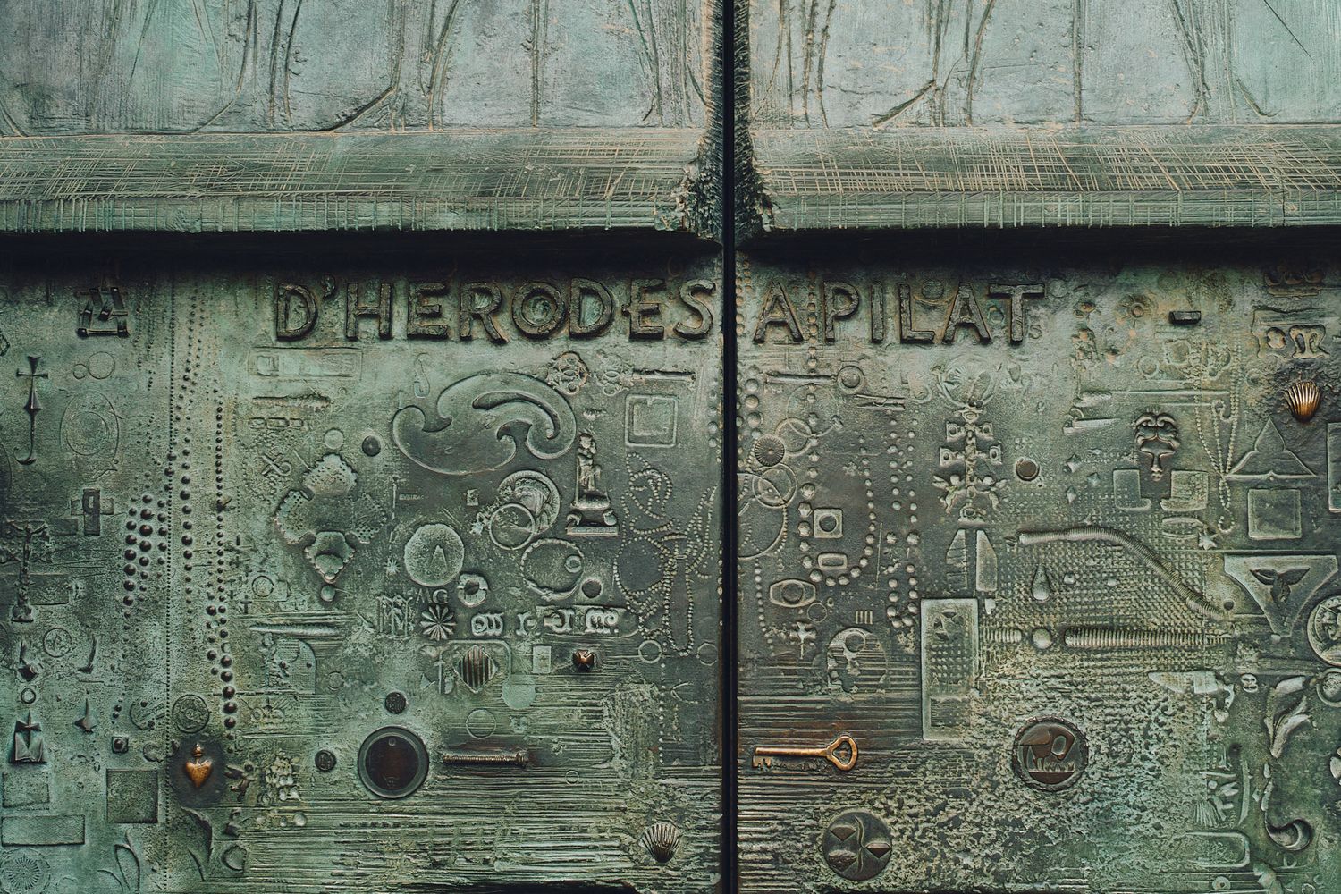 Ancient bronze door with inscription D HERODES A PILAT, featuring intricate relief carvings of symbols and figures.