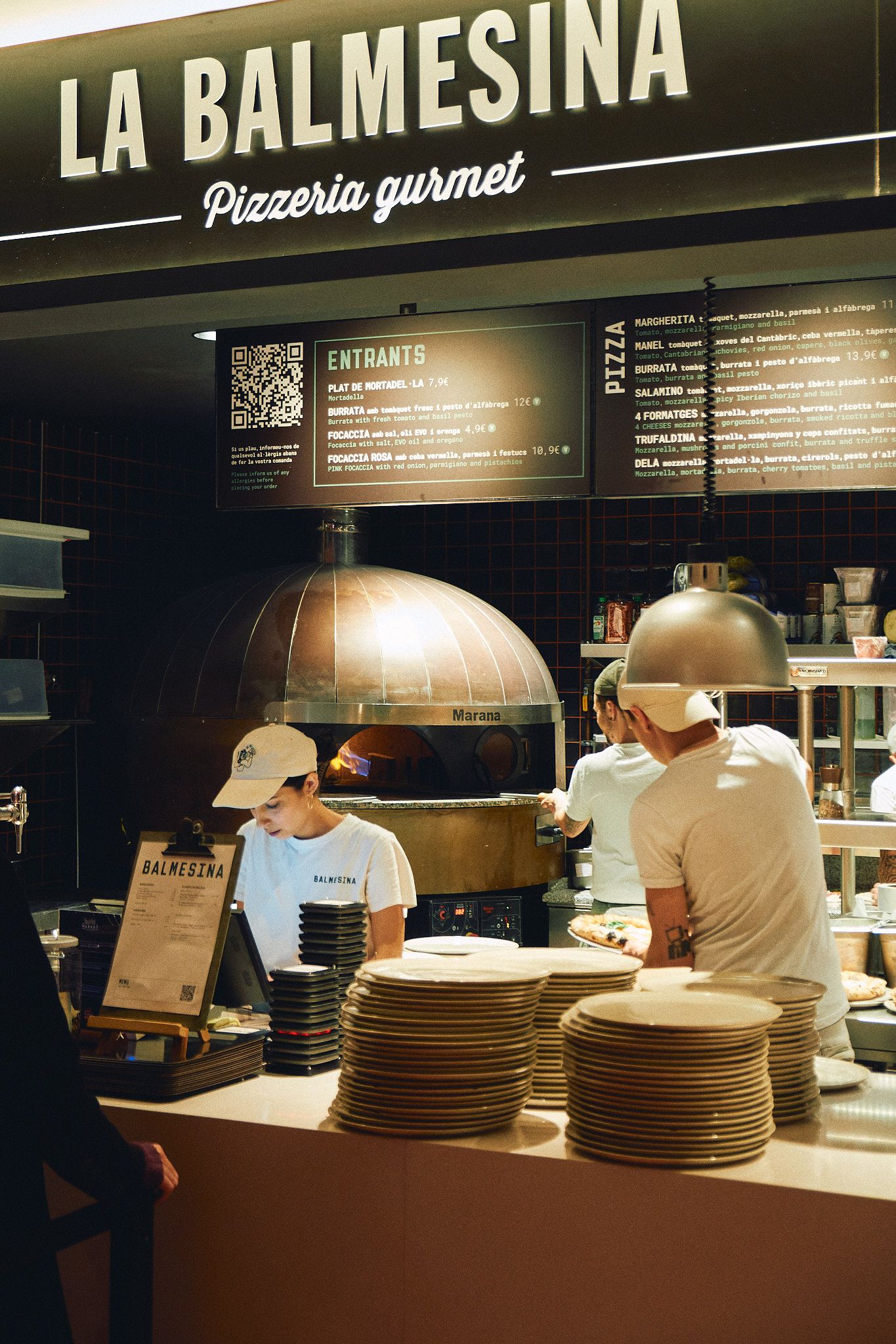 Staff preparing pizzas at La Balmesina Pizzeria Gourmet with a copper wood-fired oven and stacked plates.