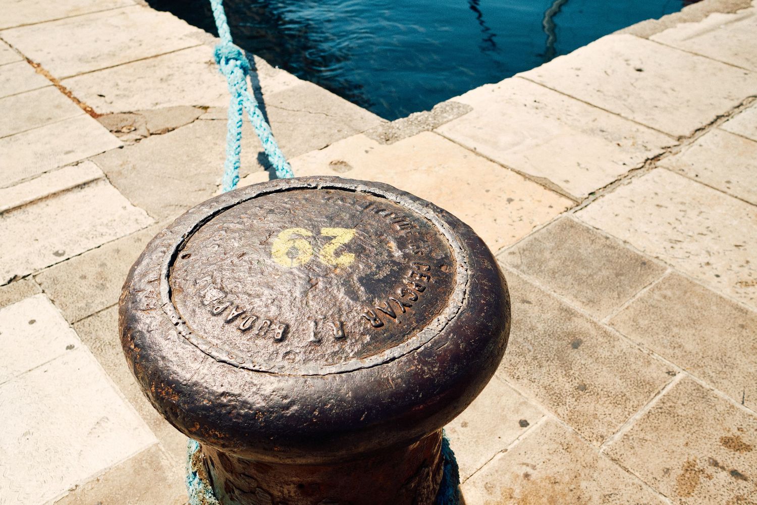 Weathered metal mooring bollard on stone dock with turquoise rope tied around it.