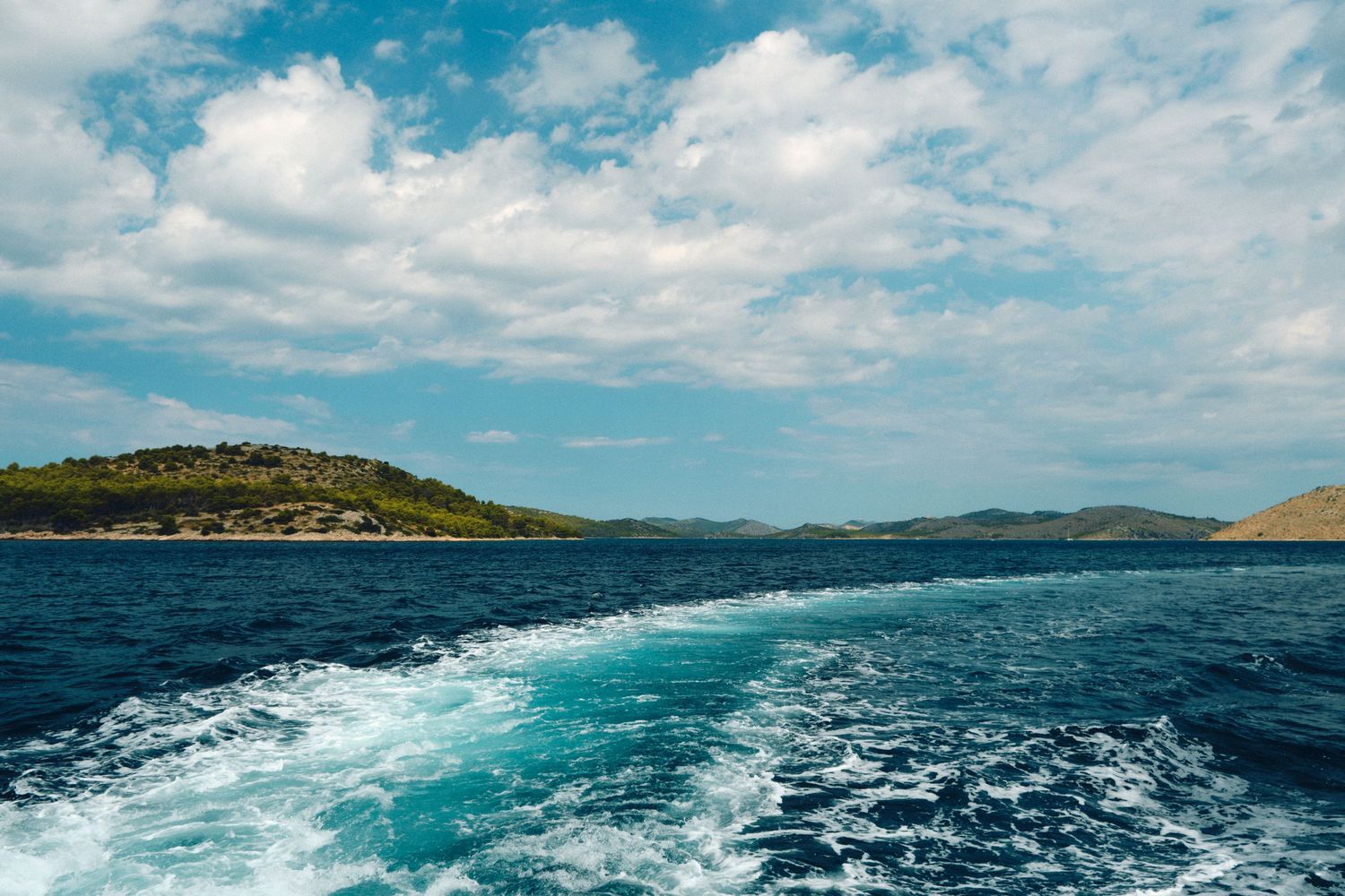 Wake patterns in turquoise Mediterranean waters with islands and blue sky in the background.