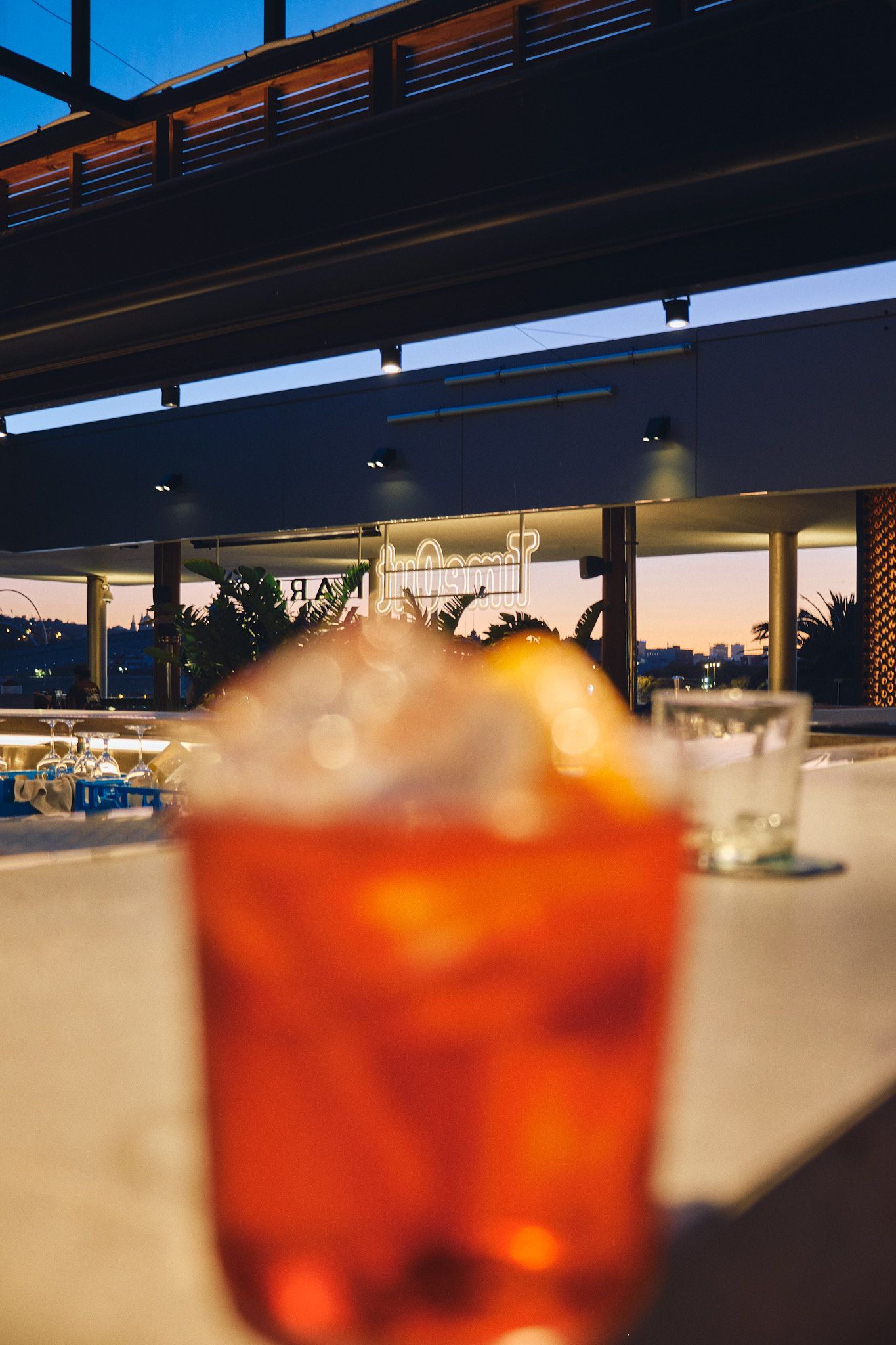 A blurred Aperol Spritz cocktail on a rooftop bar counter at dusk with city lights in the background.