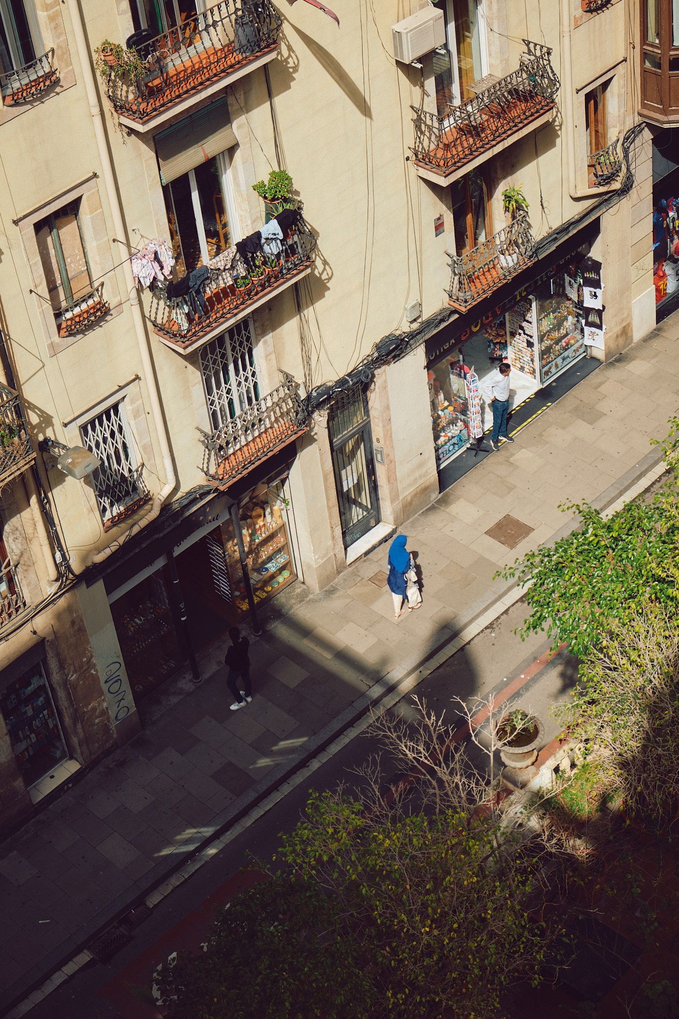 Aerial view of a European street with pedestrians walking past a shop and ornate balconied building in warm sunlight.
