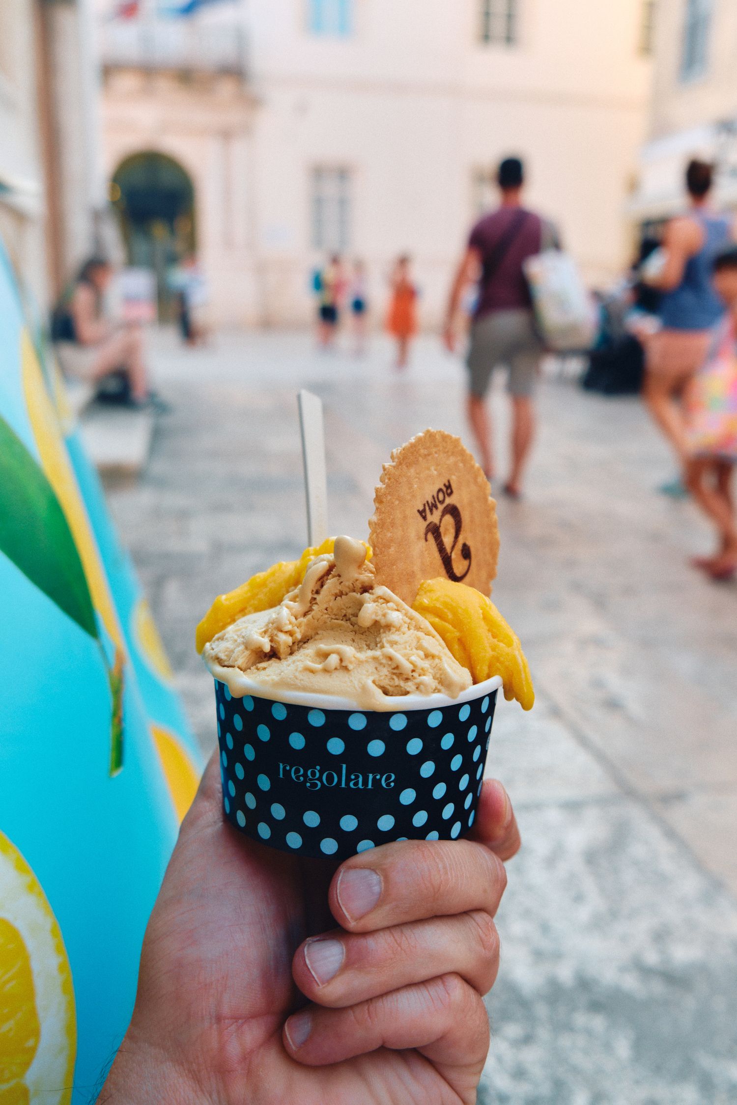 Hand holding artisanal ice cream cone with wafer cookie in a polka dot cup on cobblestone street.