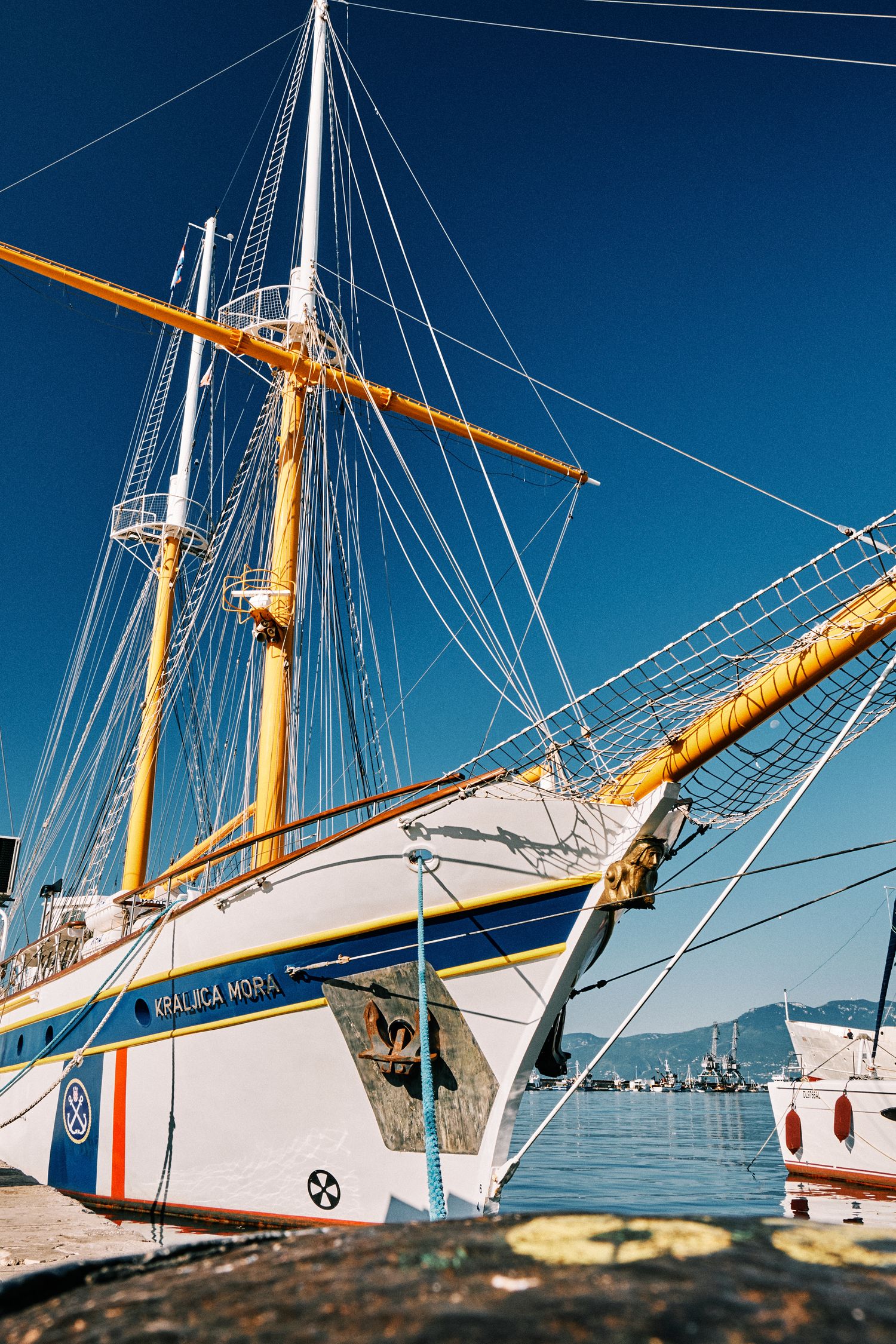 White and blue sailboat with tall masts docked at marina against clear blue sky.