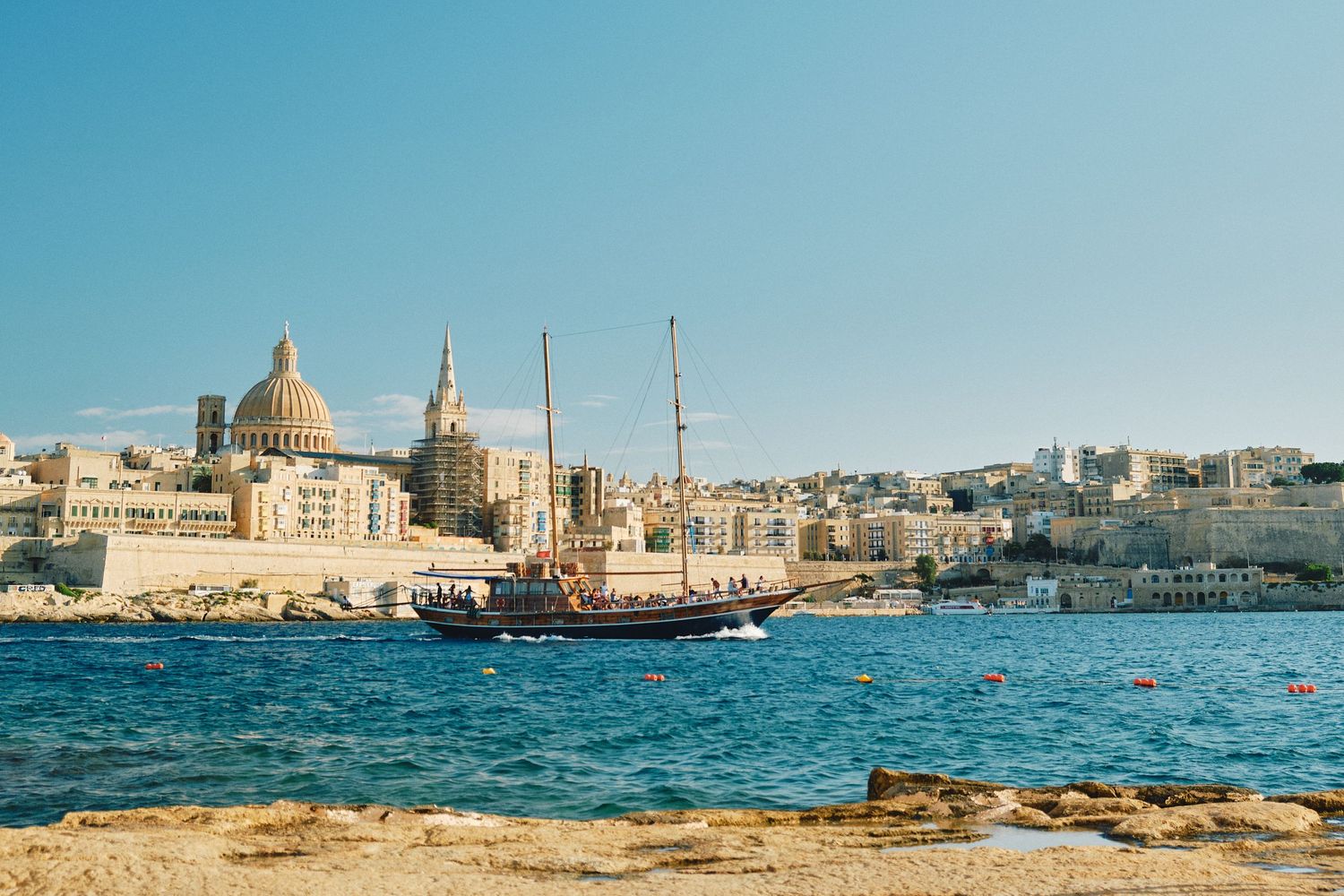 Historic Valletta waterfront with domed cathedral and traditional sailing ship on blue Mediterranean waters.