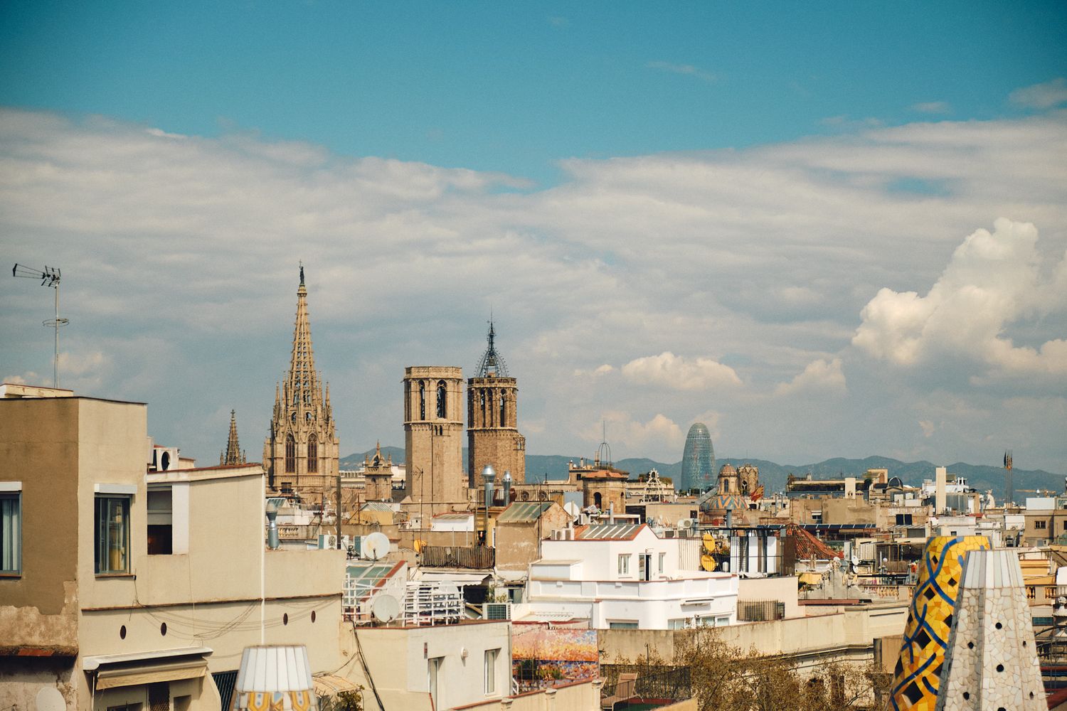 Panoramic view of Barcelona skyline featuring Gothic cathedral spires and modern towers under a partly cloudy blue sky.