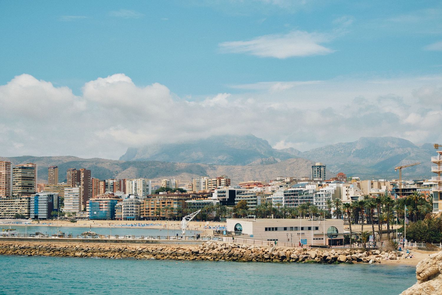 Mediterranean coastal cityscape with modern buildings and mountains visible in the background.