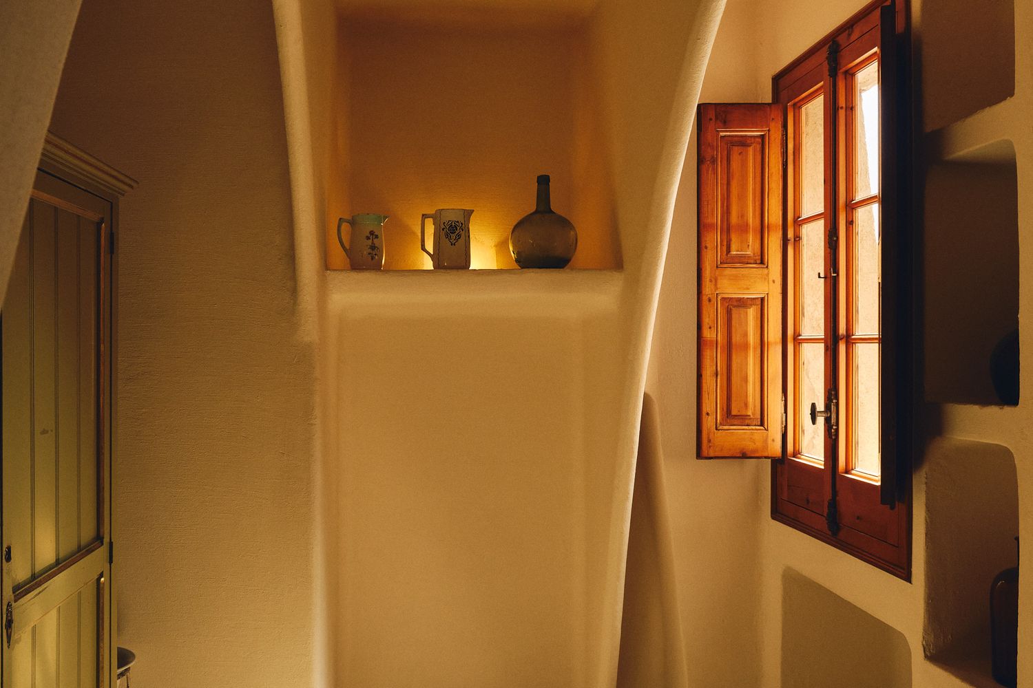 Warm Santorini interior with arched white walls, wooden window shutters, and decorative shelf with vases.