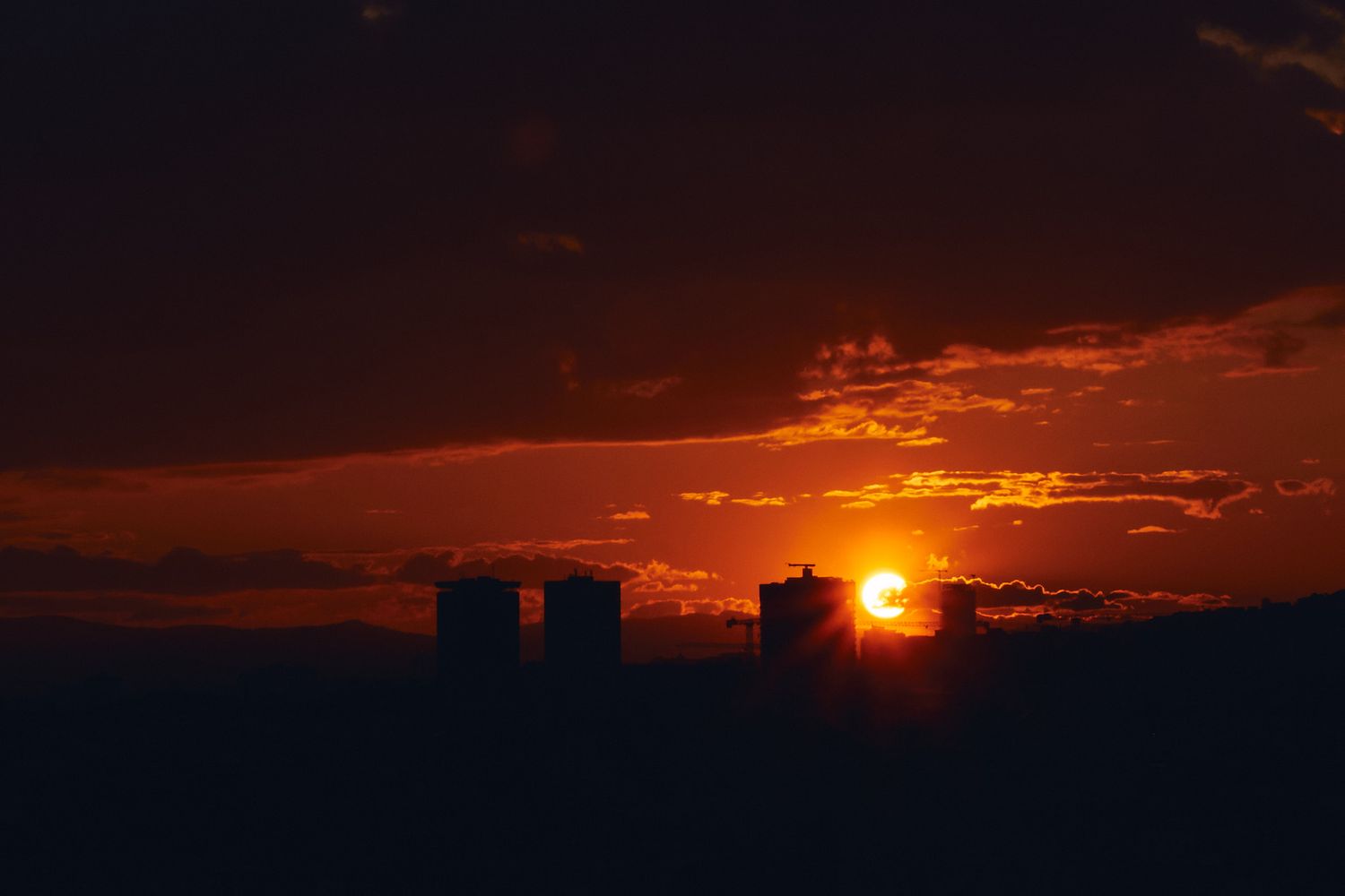 Dramatic urban sunset silhouette with glowing orange sun behind city buildings against dark cloudy sky.