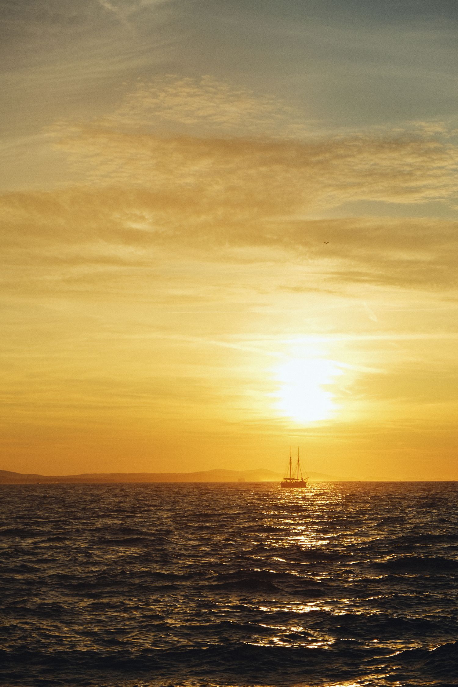 Sailboat silhouetted against dramatic golden sunset over rippling ocean waters.