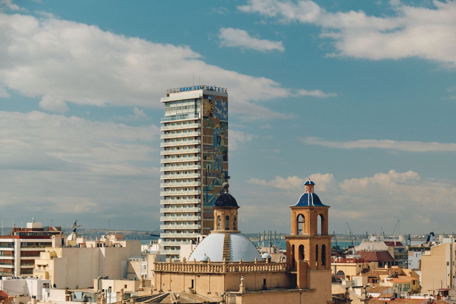 Modern high-rise building contrasts with historic church towers against bright blue sky.