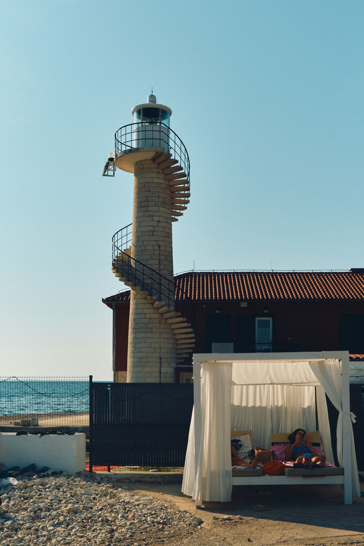 A lighthouse with spiral staircase and white canopy lounge area by the Mediterranean Sea on a sunny day.