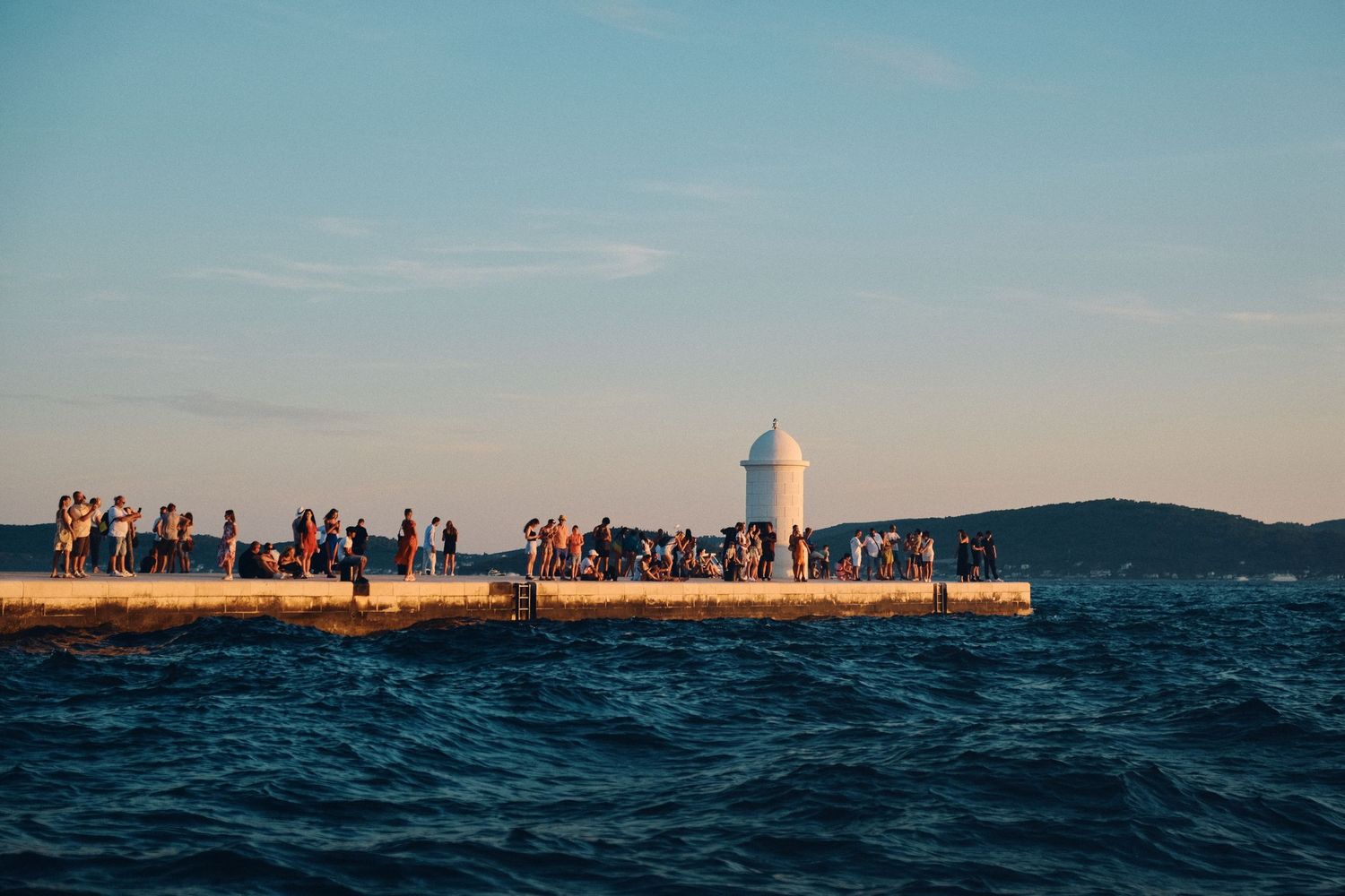 People gathered on coastal breakwater at dusk with lighthouse in distance.