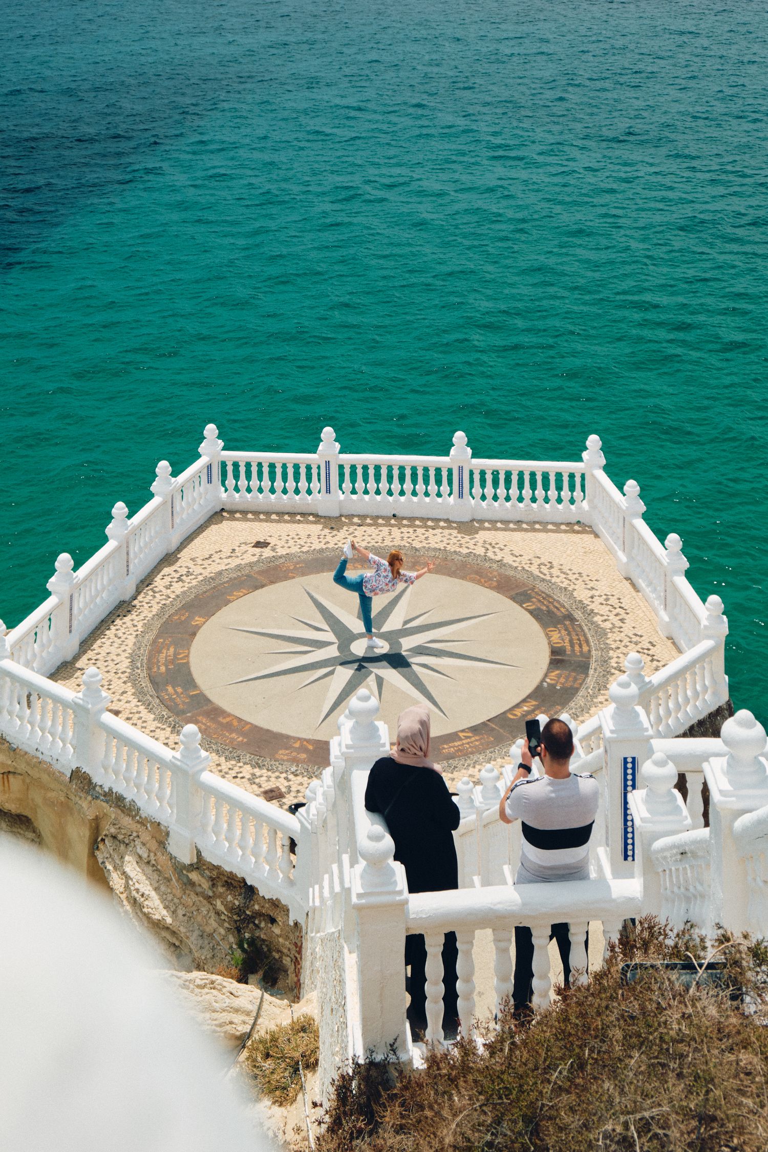 Circular white balcony overlook with compass rose design floor pattern above turquoise Mediterranean waters.
