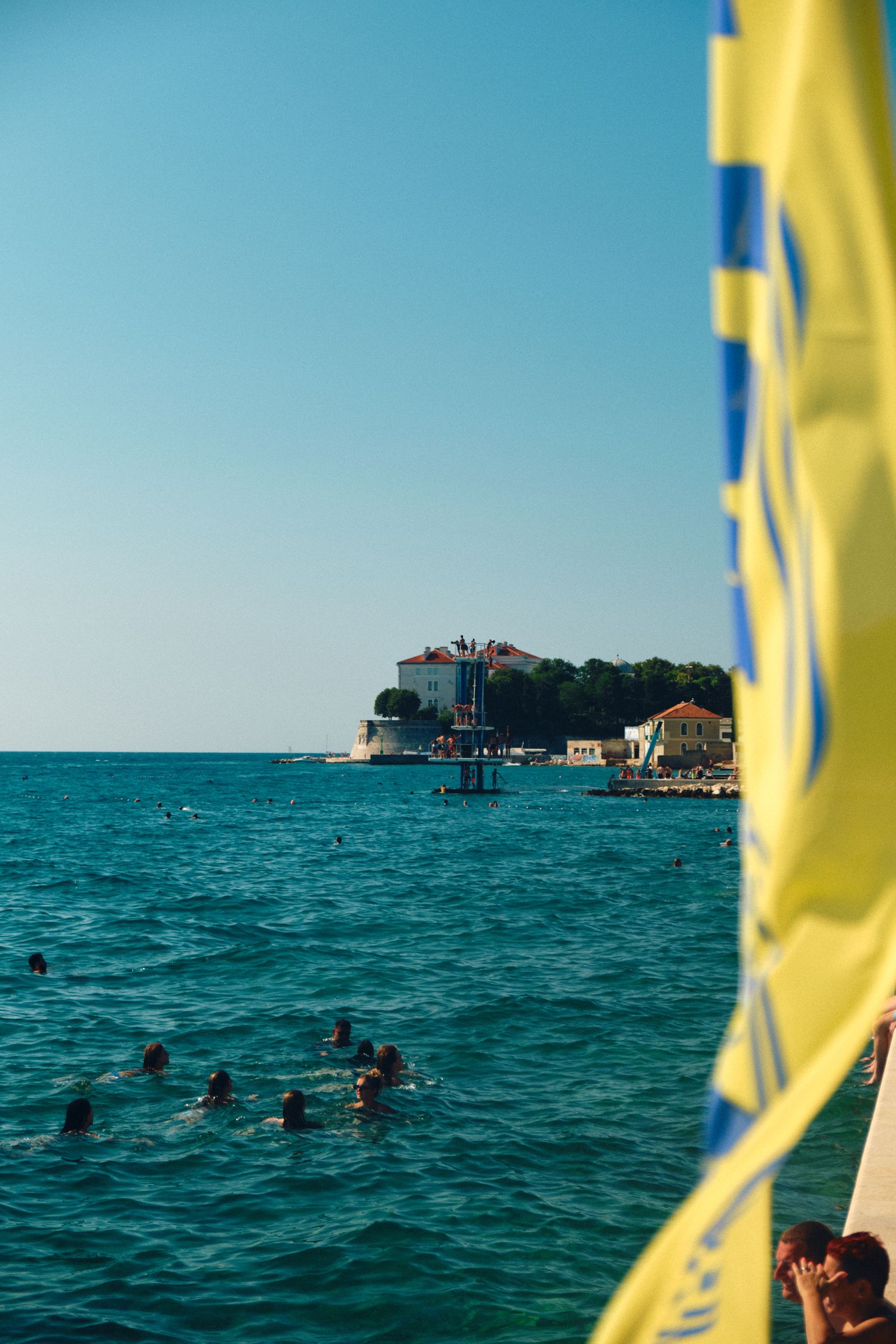 Swimmers enjoying crystal clear turquoise waters near coastal buildings on a sunny Mediterranean day.