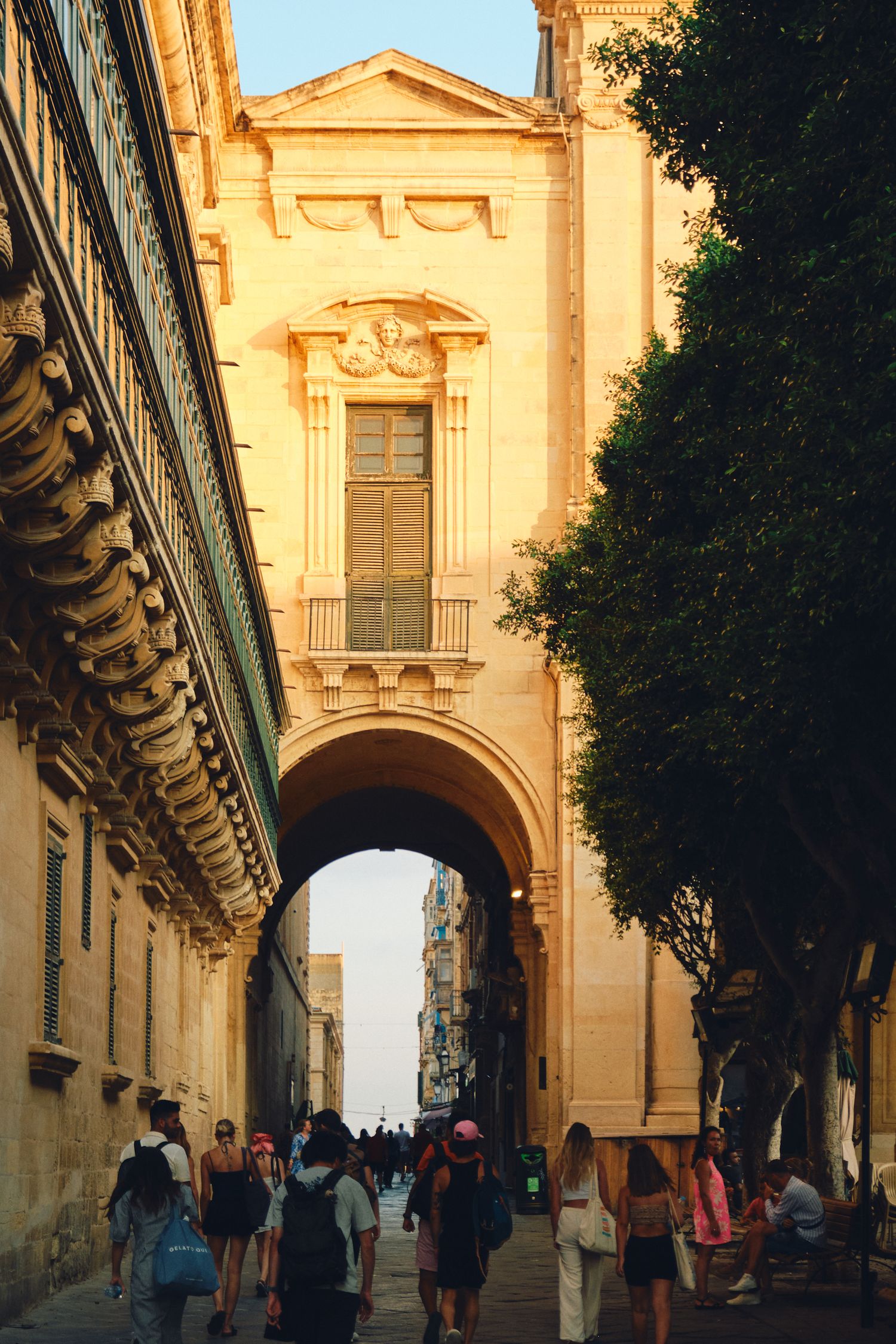 Historic archway passage with tourists walking through golden-lit limestone architecture.