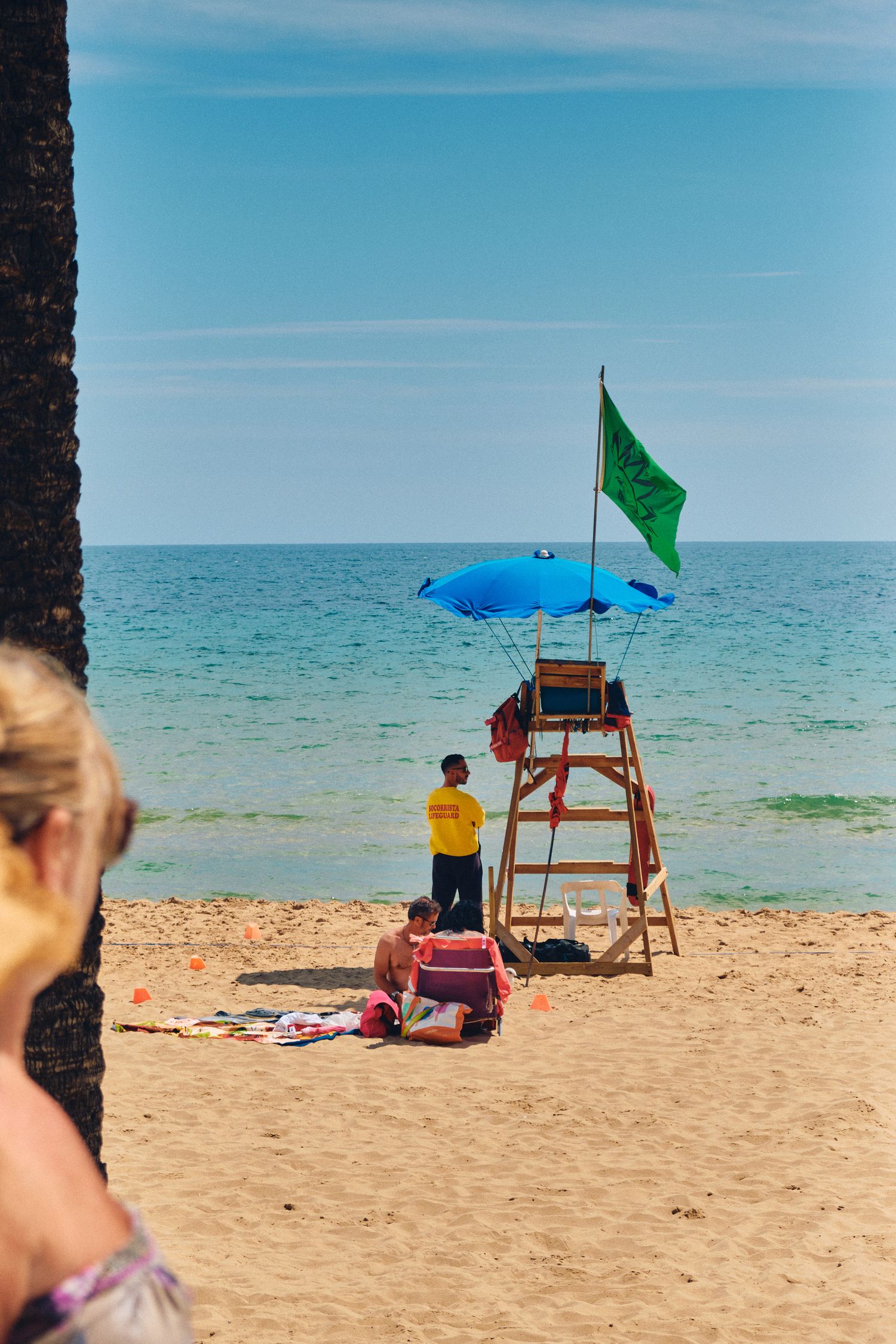 Lifeguard station with green flag and blue umbrella on sandy beach overlooking turquoise ocean.