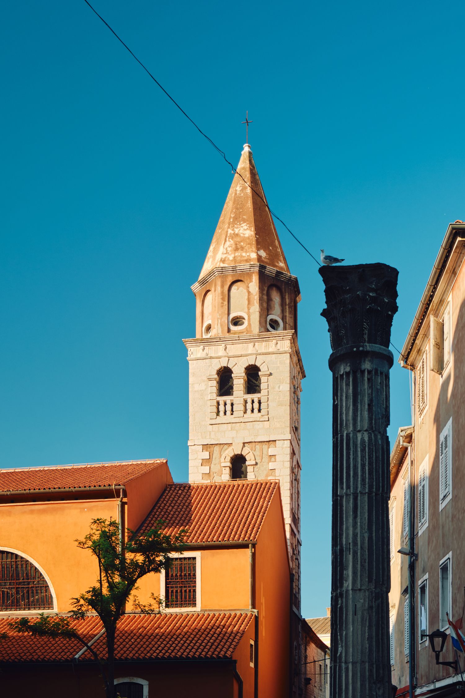 Church bell tower with terracotta roof and ancient Roman column against bright blue sky.