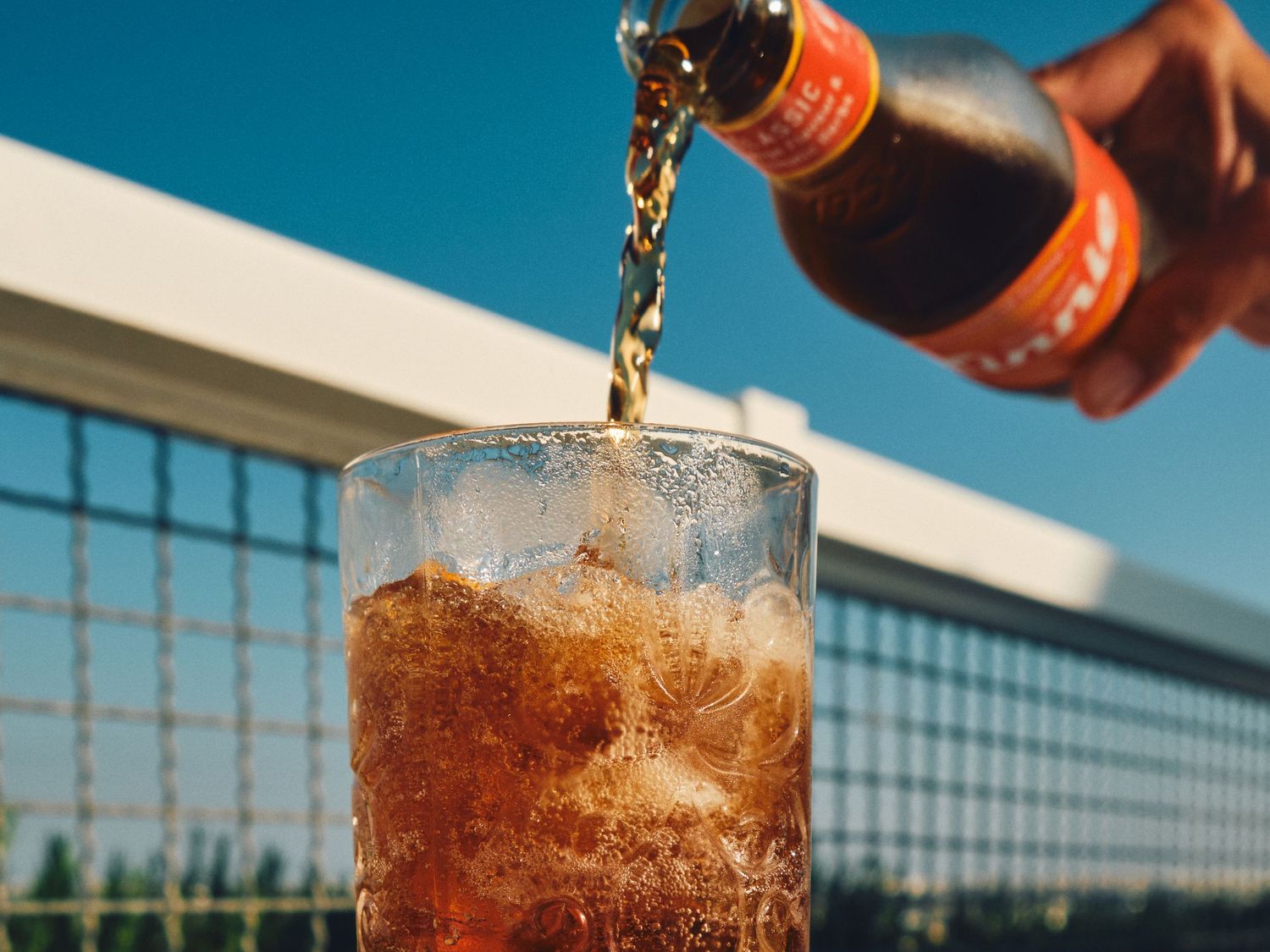 Carbonated beverage being poured into an ice-filled glass against a blue sky.