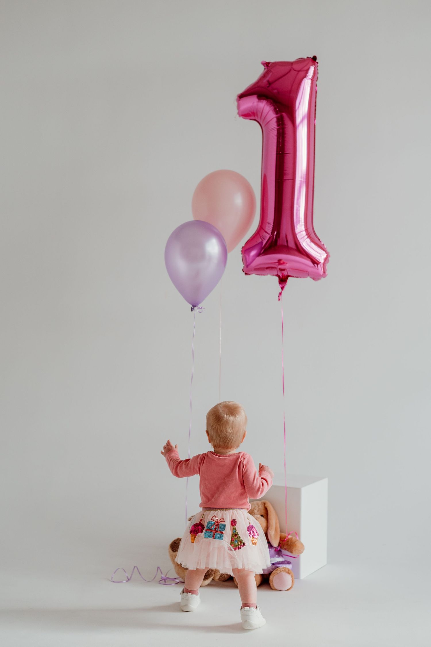 Small child in pink shirt and patterned skirt sits by pink number one balloon and pastel decorations for first birthday.