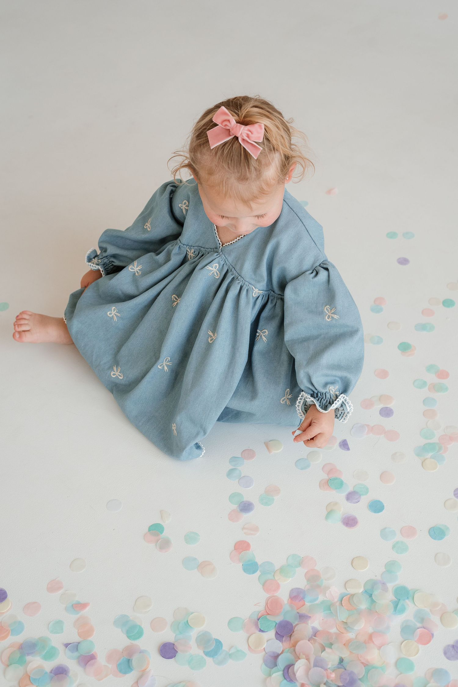 A baby in a blue dress lies on white paper surrounded by pastel pink, blue, and purple confetti dots.