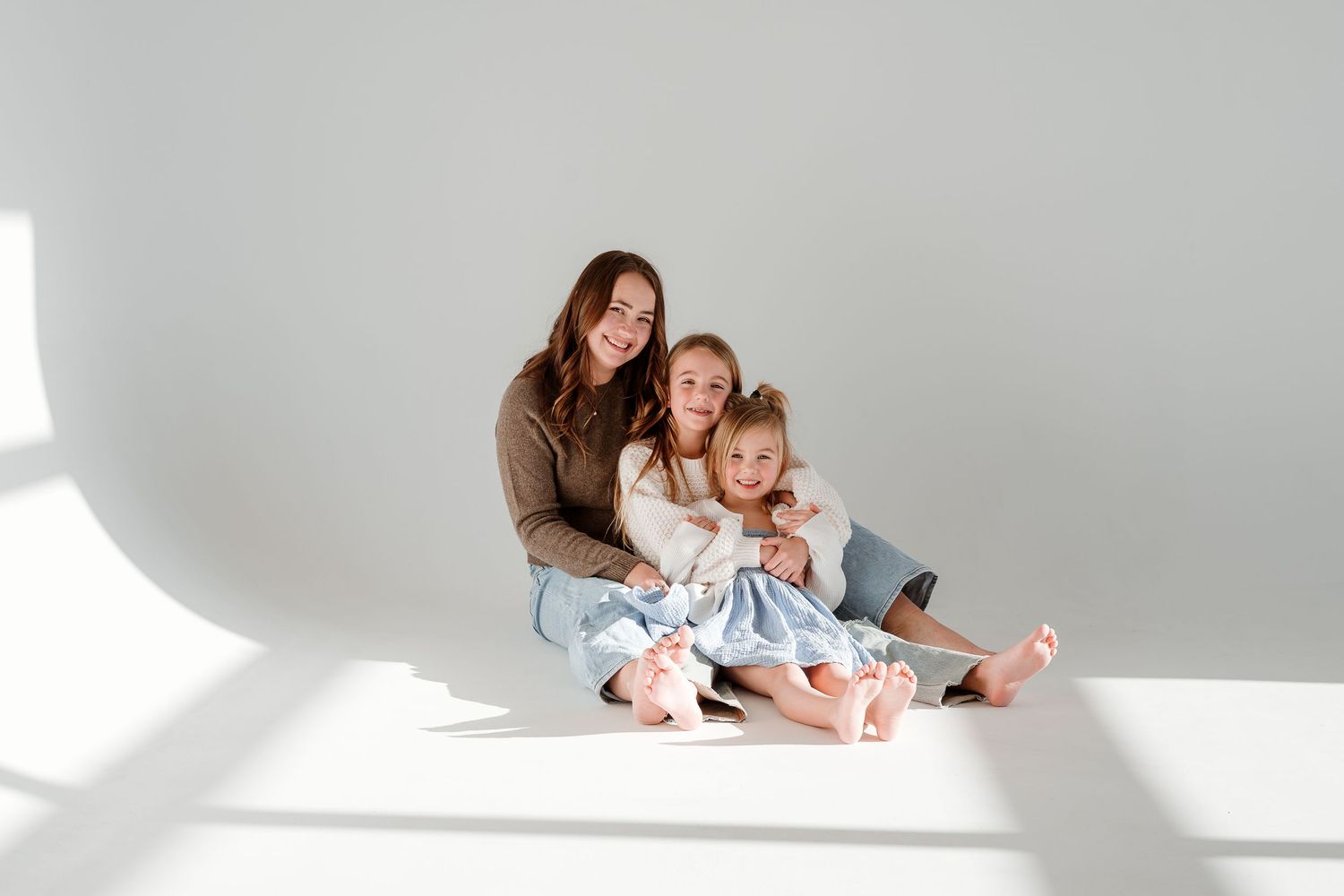 Mother and child in white outfits sitting together in minimalist studio setting with soft natural lighting.