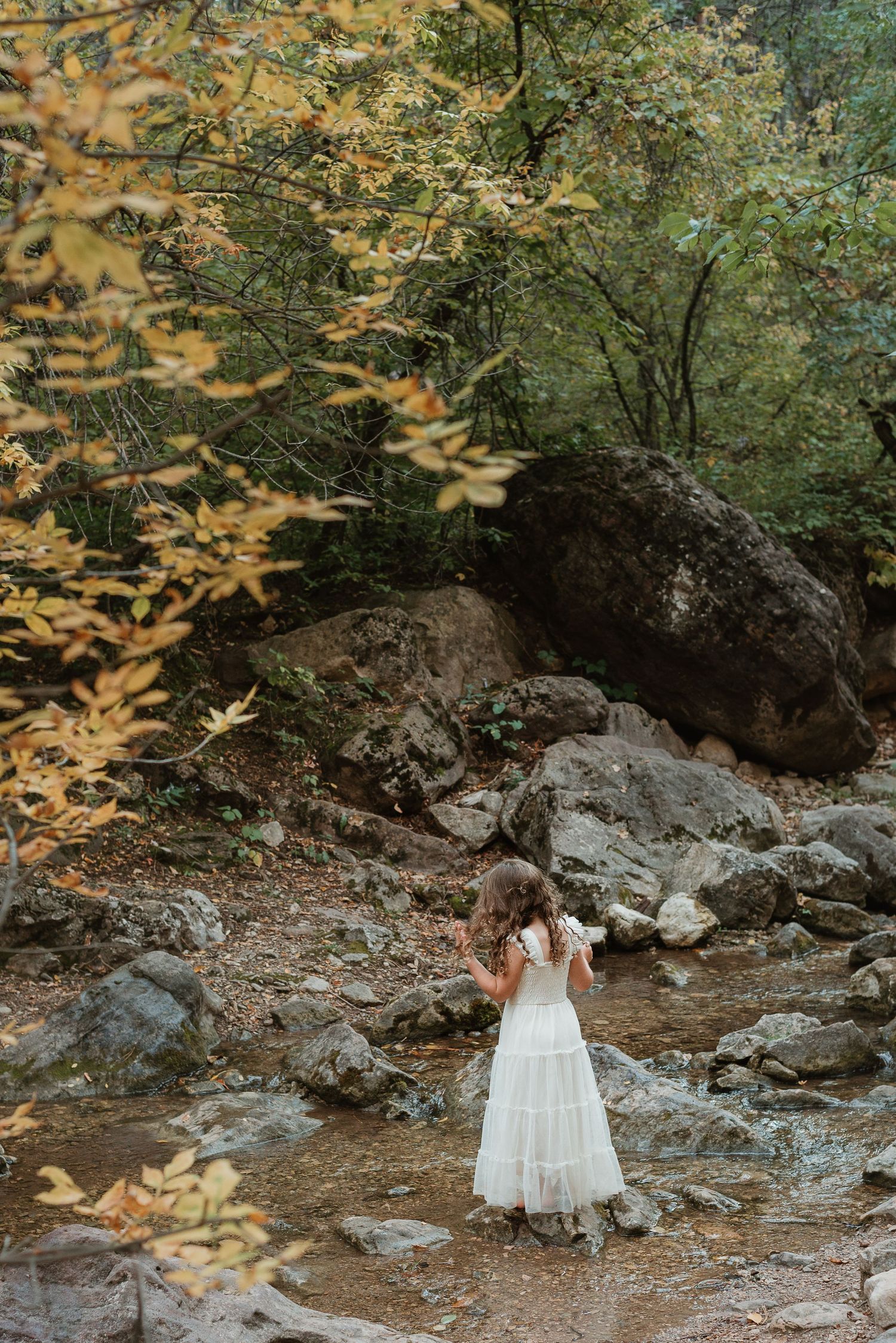 A small child in a white dress explores a rocky creek bed surrounded by autumn foliage and large boulders.