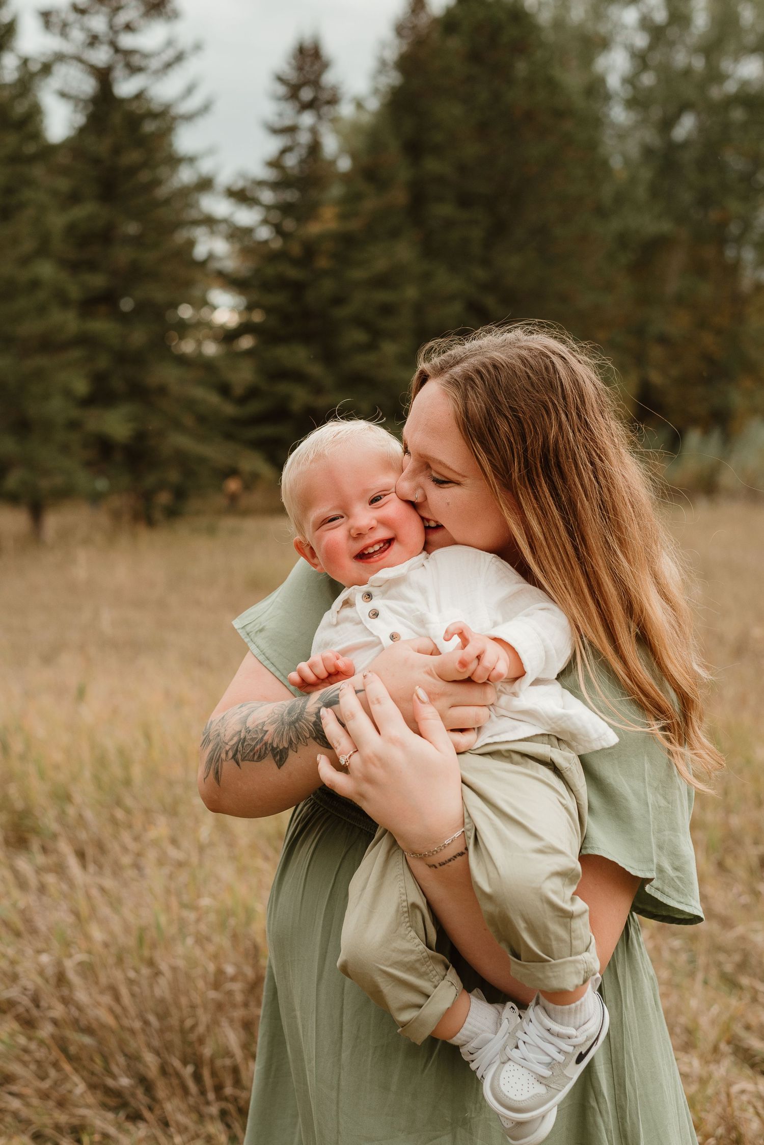A tender moment captured in a field as a mother in sage green embraces and kisses a joyful baby in white.