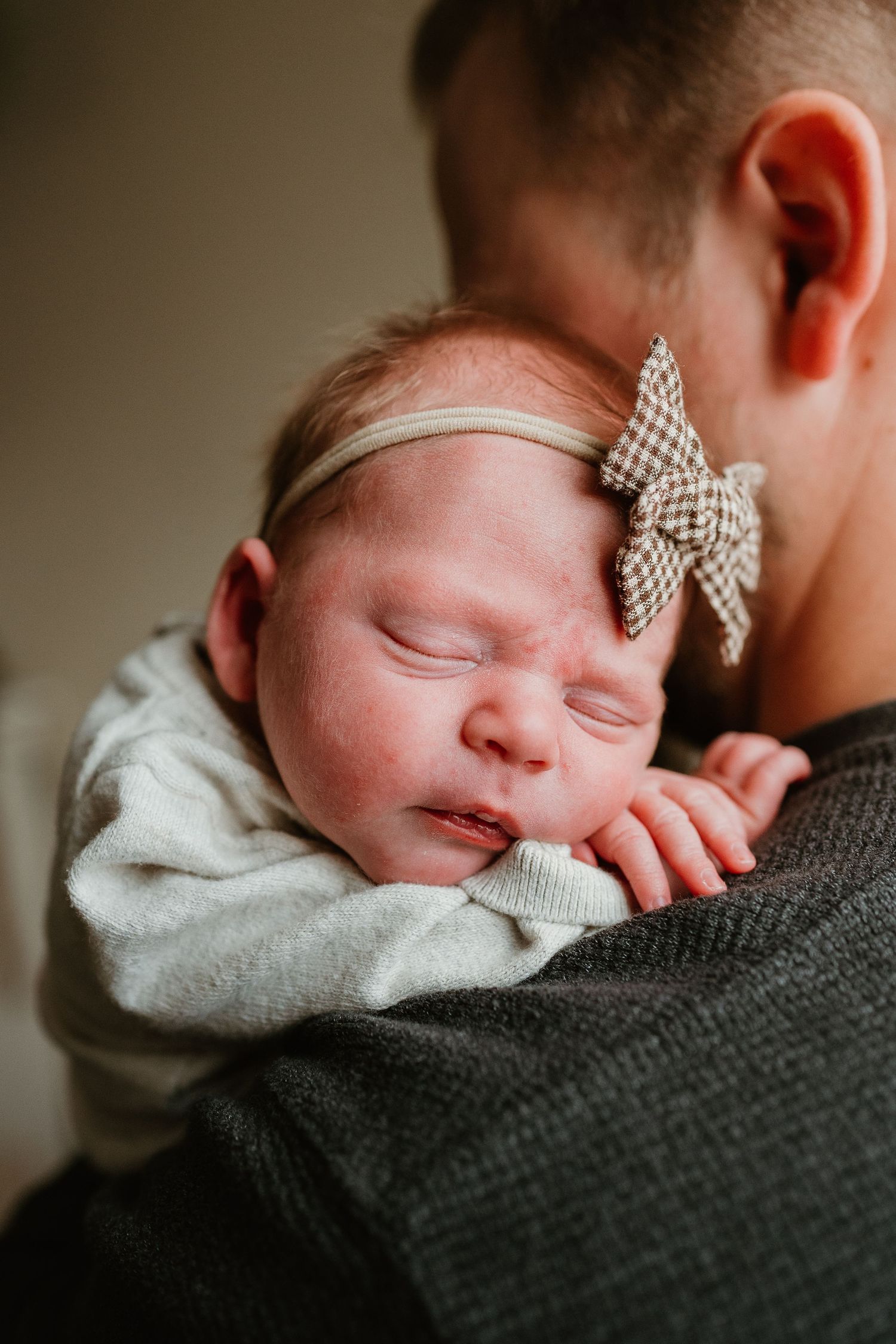 A sleeping newborn baby wearing a checkered bow headband rests peacefully against someone's shoulder in a grey sweater.