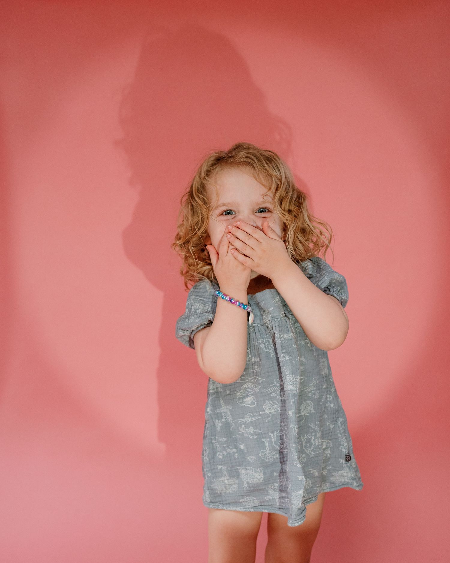 A child in a gray dress covers their mouth with both hands while standing against a pink background.