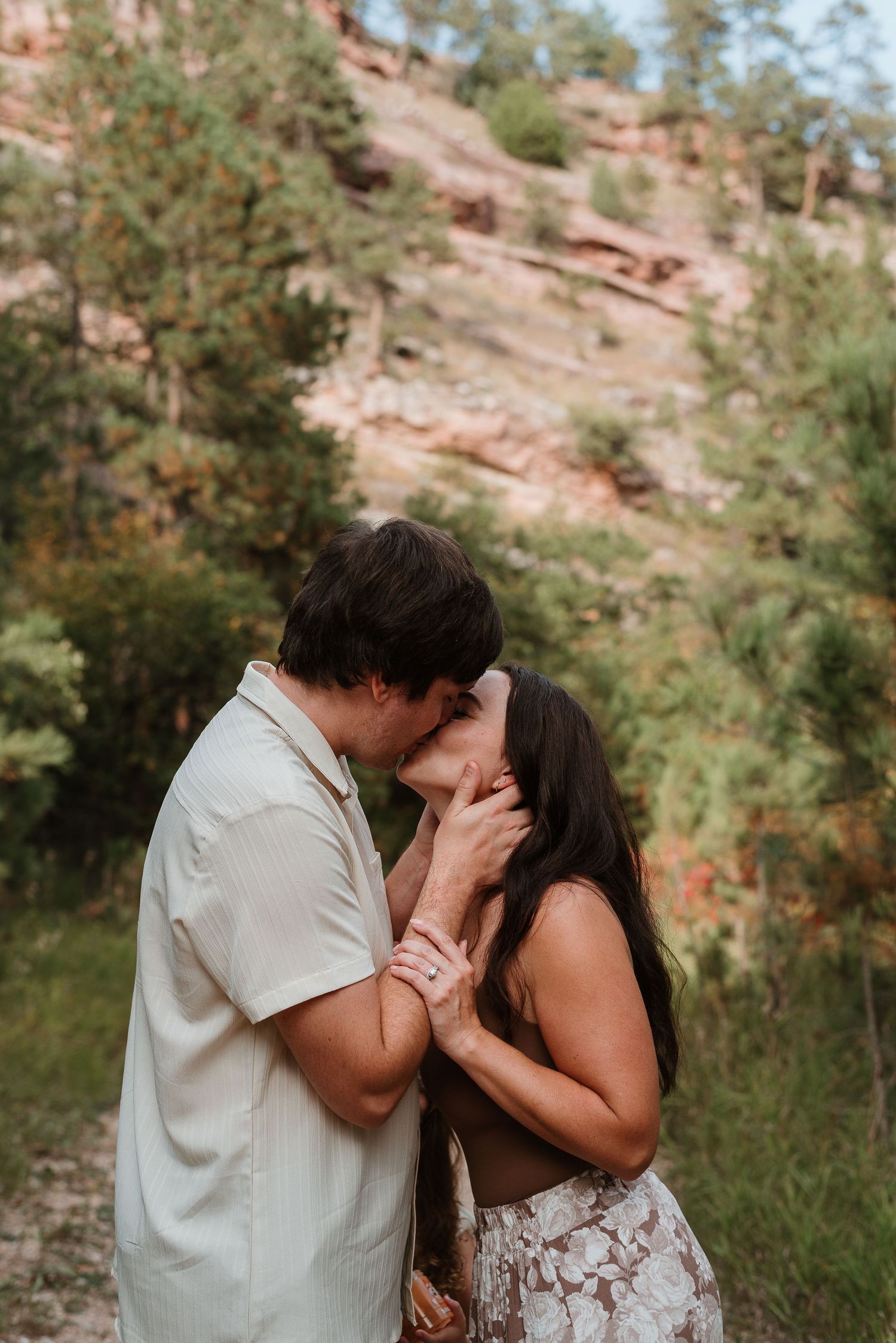A romantic couple shares tender moments in a natural outdoor setting with red rock formations and greenery.