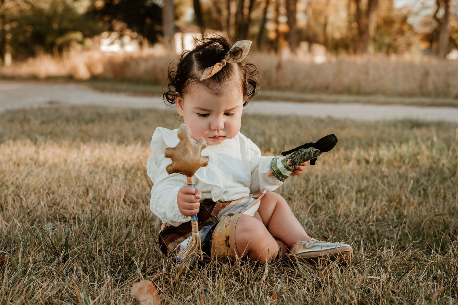 Small child in white clothing sits in grass playing with toys during golden hour.