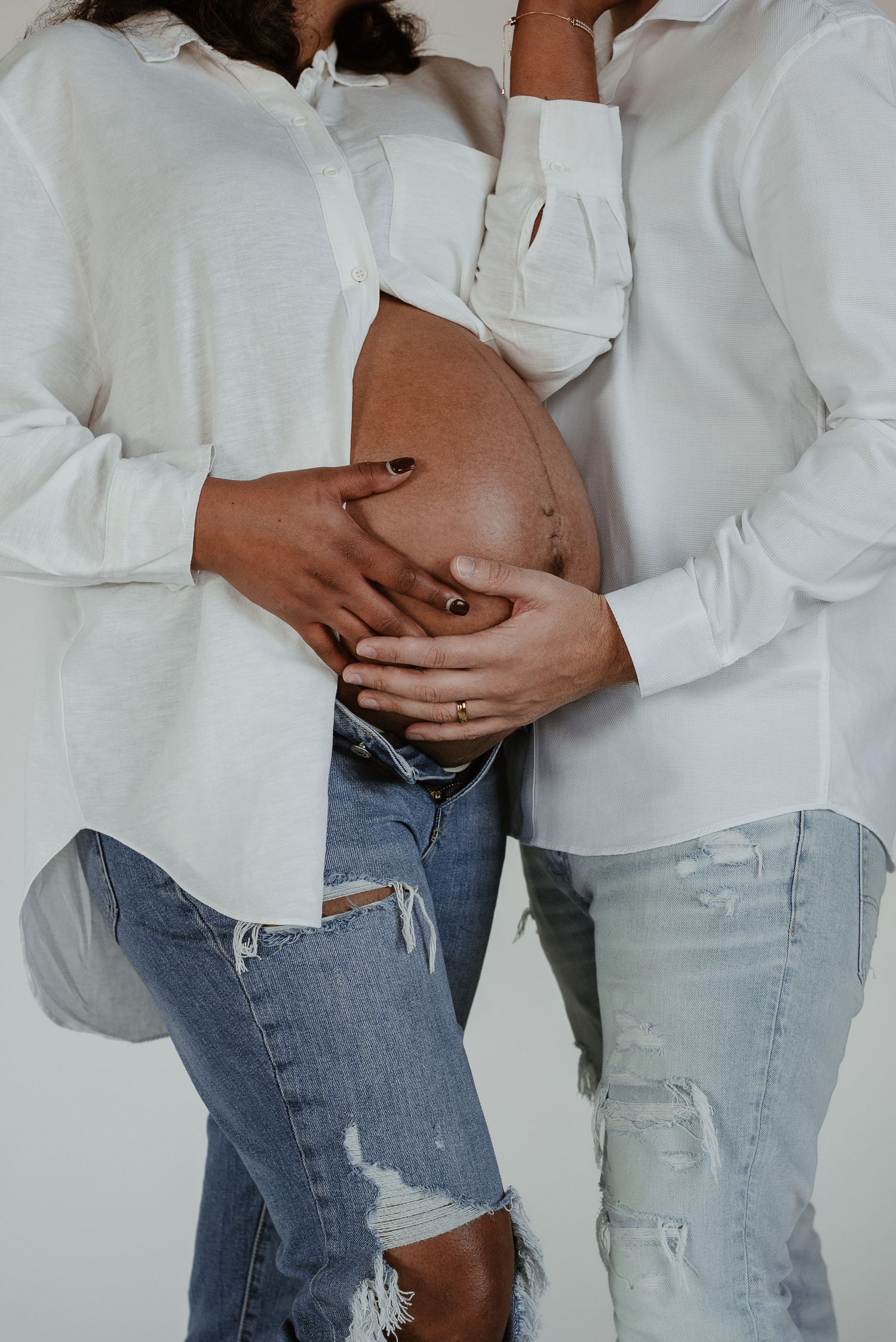 Intimate maternity photo showing couple in white shirts and distressed jeans embracing pregnant belly.