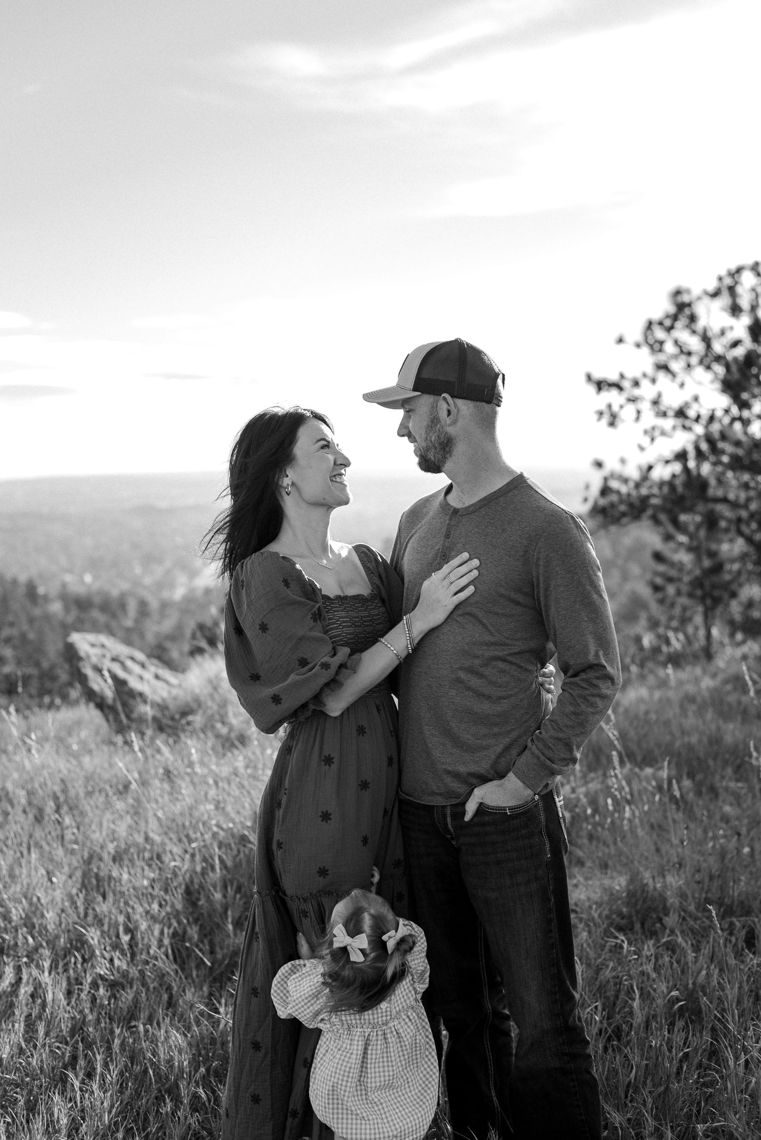 A couple shares a romantic moment together on a hilltop overlooking a scenic landscape in black and white photography.