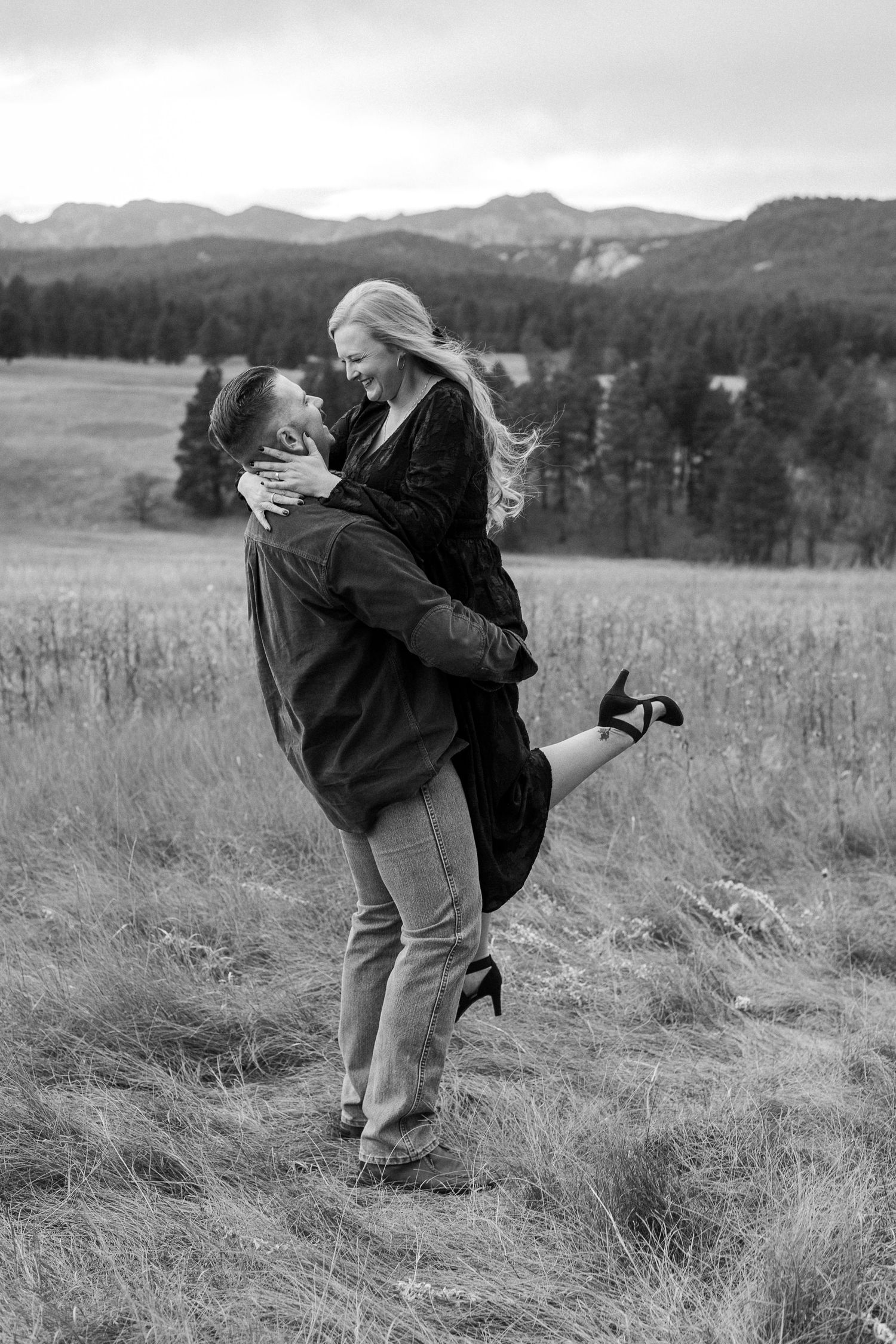 A romantic black and white photo of a couple embracing in a mountain meadow with evergreen forests in the background.
