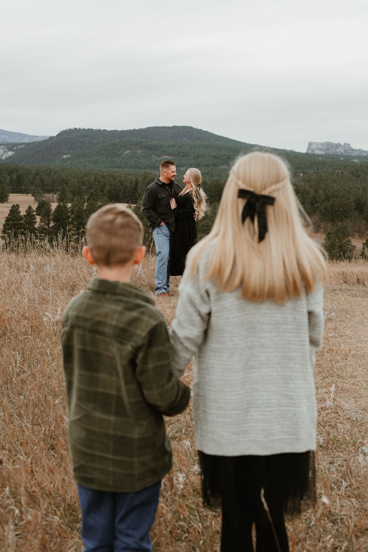 A couple and child walking through a golden field with mountains and forest in the background.