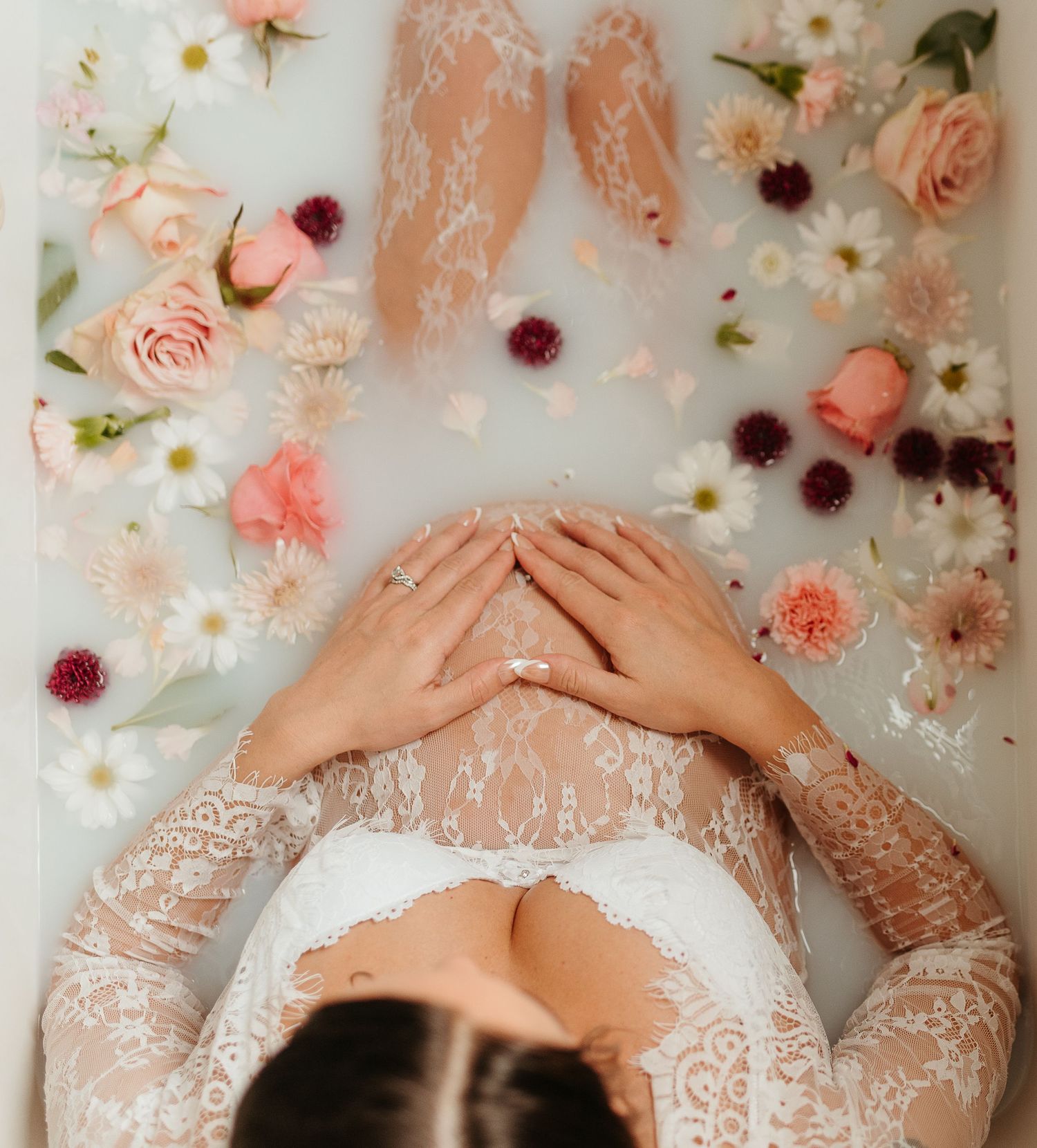 A milk bath maternity photo with delicate lace and scattered pink and burgundy flowers creating a romantic atmosphere.