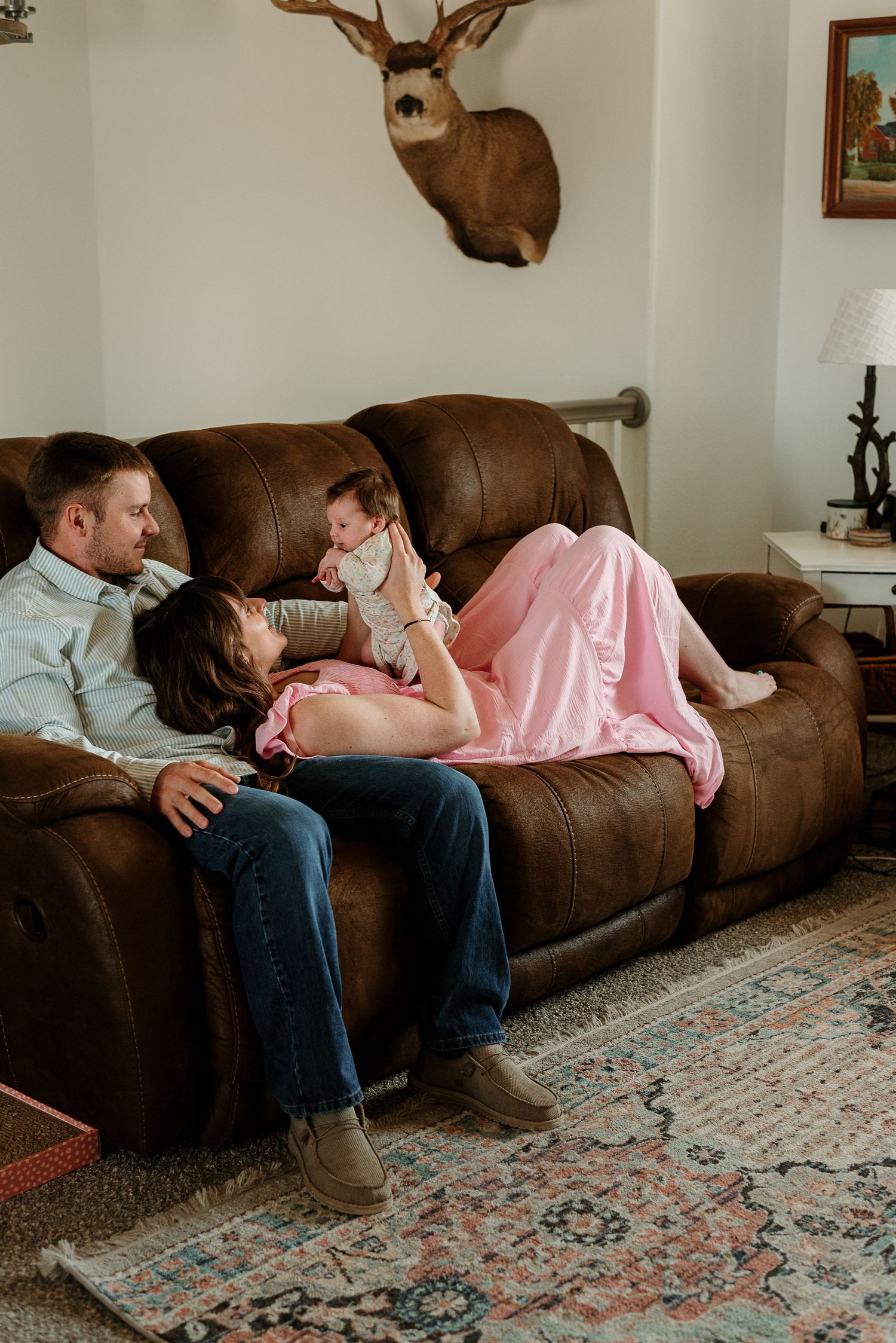 Two people relax together on a brown leather couch in a cozy living room with mounted deer antlers on the wall.