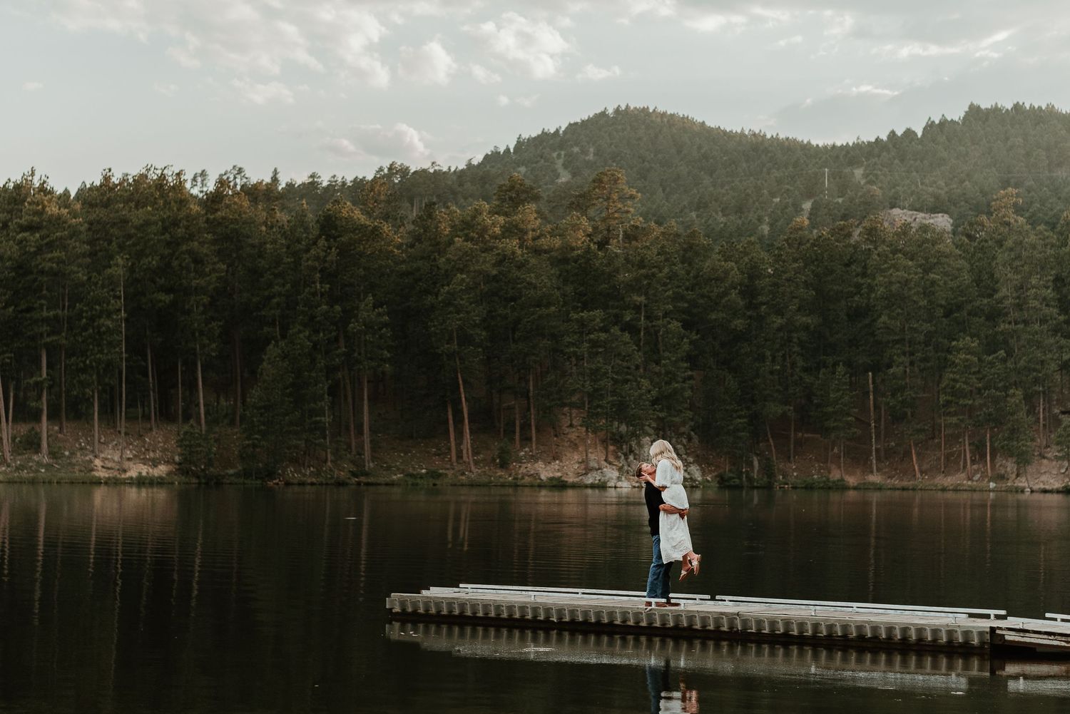 Couple standing on a wooden dock overlooking a serene mountain lake.