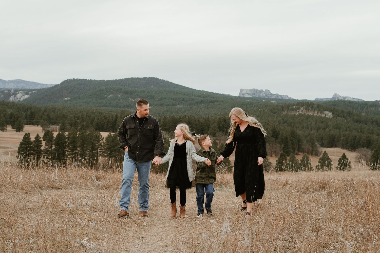 A group walks through a scenic mountain meadow with evergreen trees and rolling hills in the background.