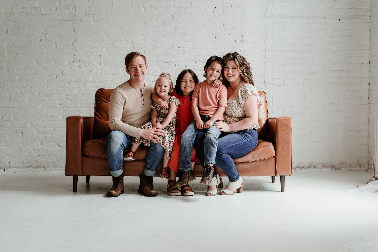 Family lounges together on a brown leather couch in a minimalist white studio setting.