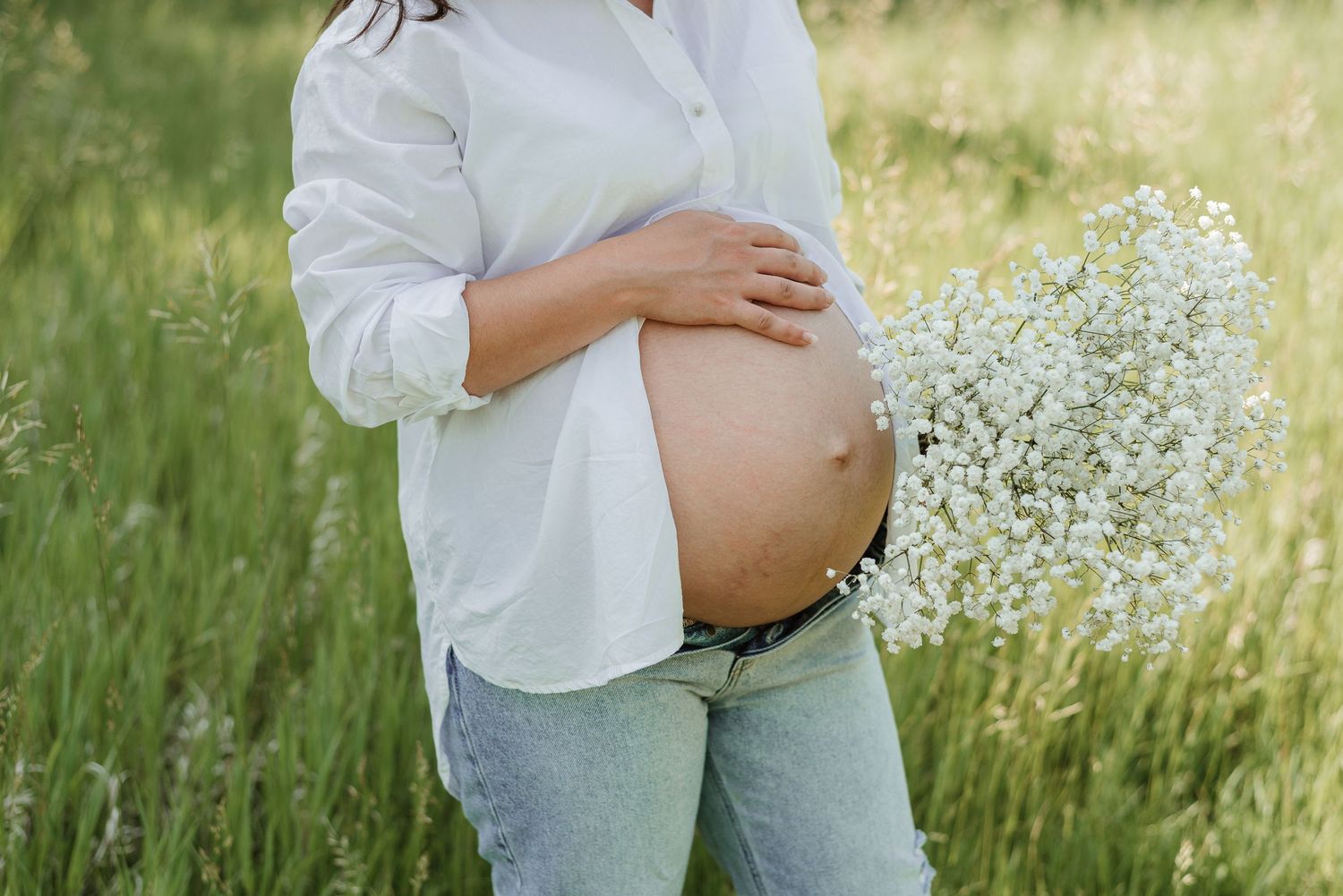 A pregnant person holds white wildflowers while standing in tall grass.
