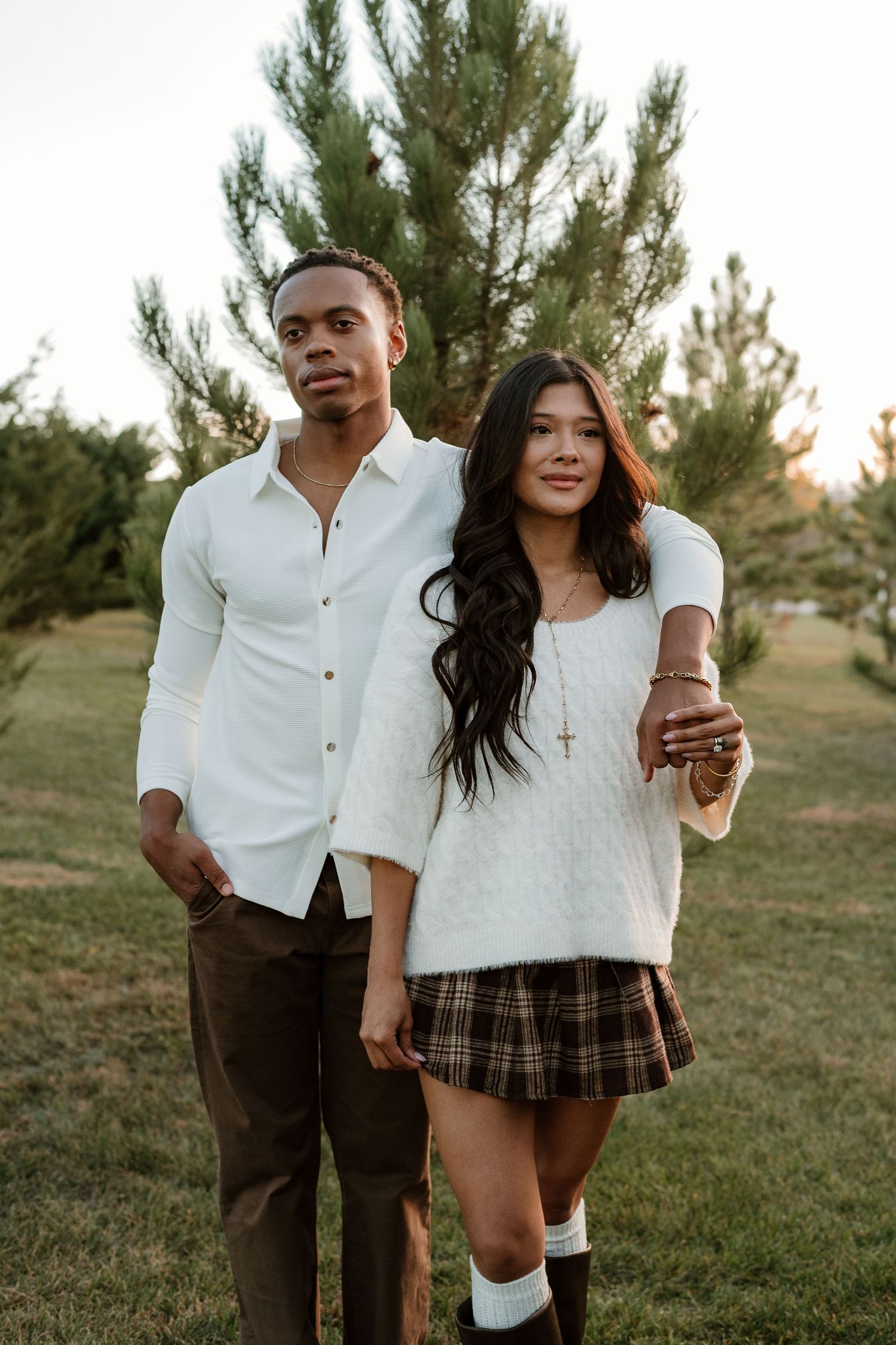Playful couple in schoolgirl-inspired outfit and white shirt posing in a grassy field with pine trees.