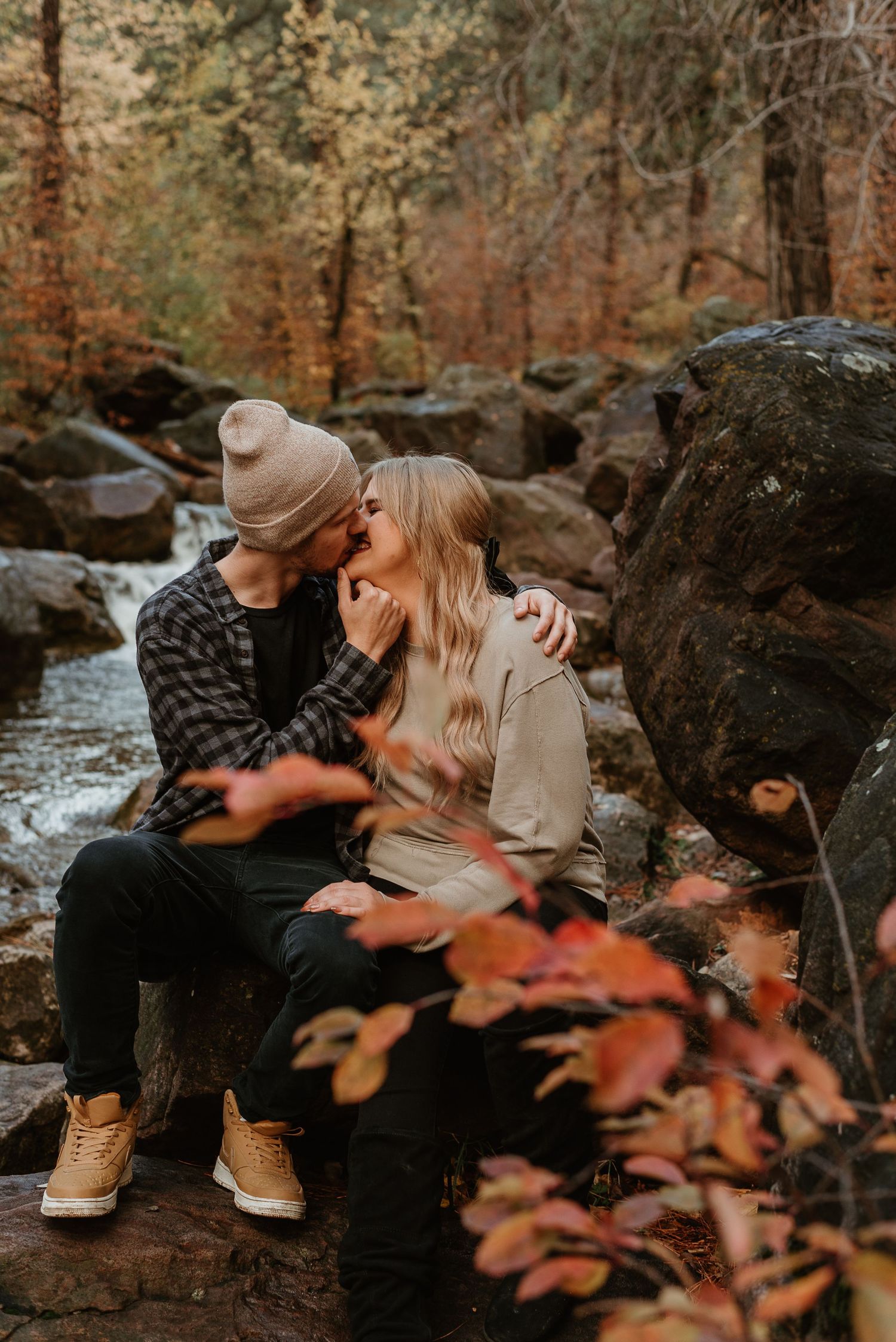 A couple sits together on rocks by a stream surrounded by autumn foliage in a romantic outdoor setting.