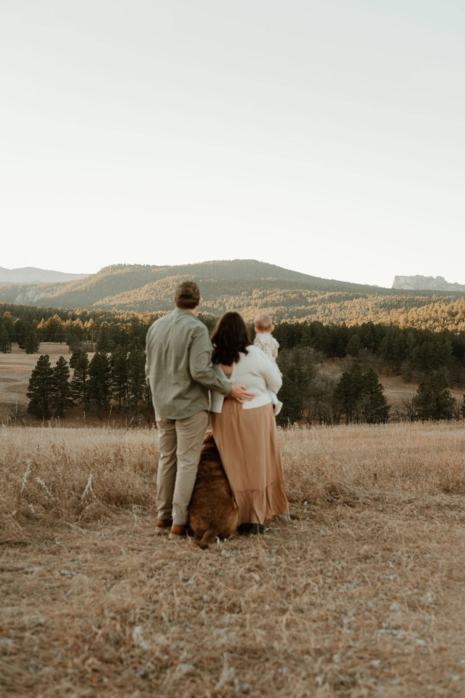 Couple sits together in a rustic field with mountains and forest in the background during golden hour.