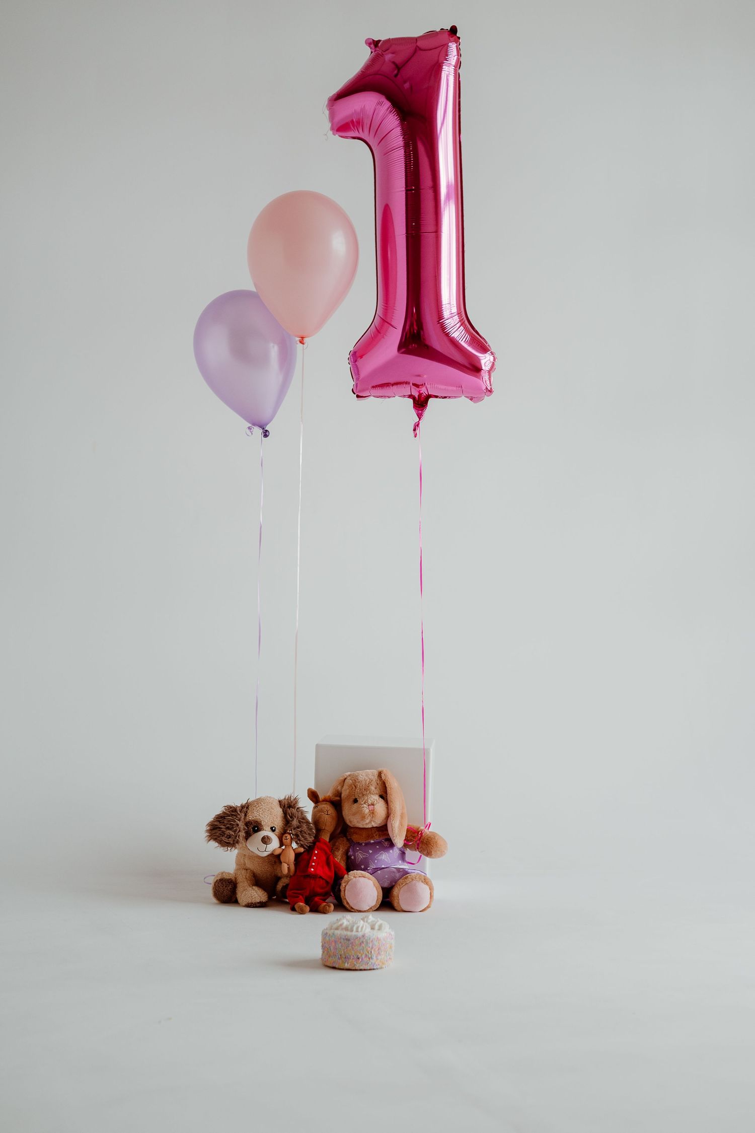 Pink number one balloon and pastel balloons float above stuffed animals and toys arranged on white surface.
